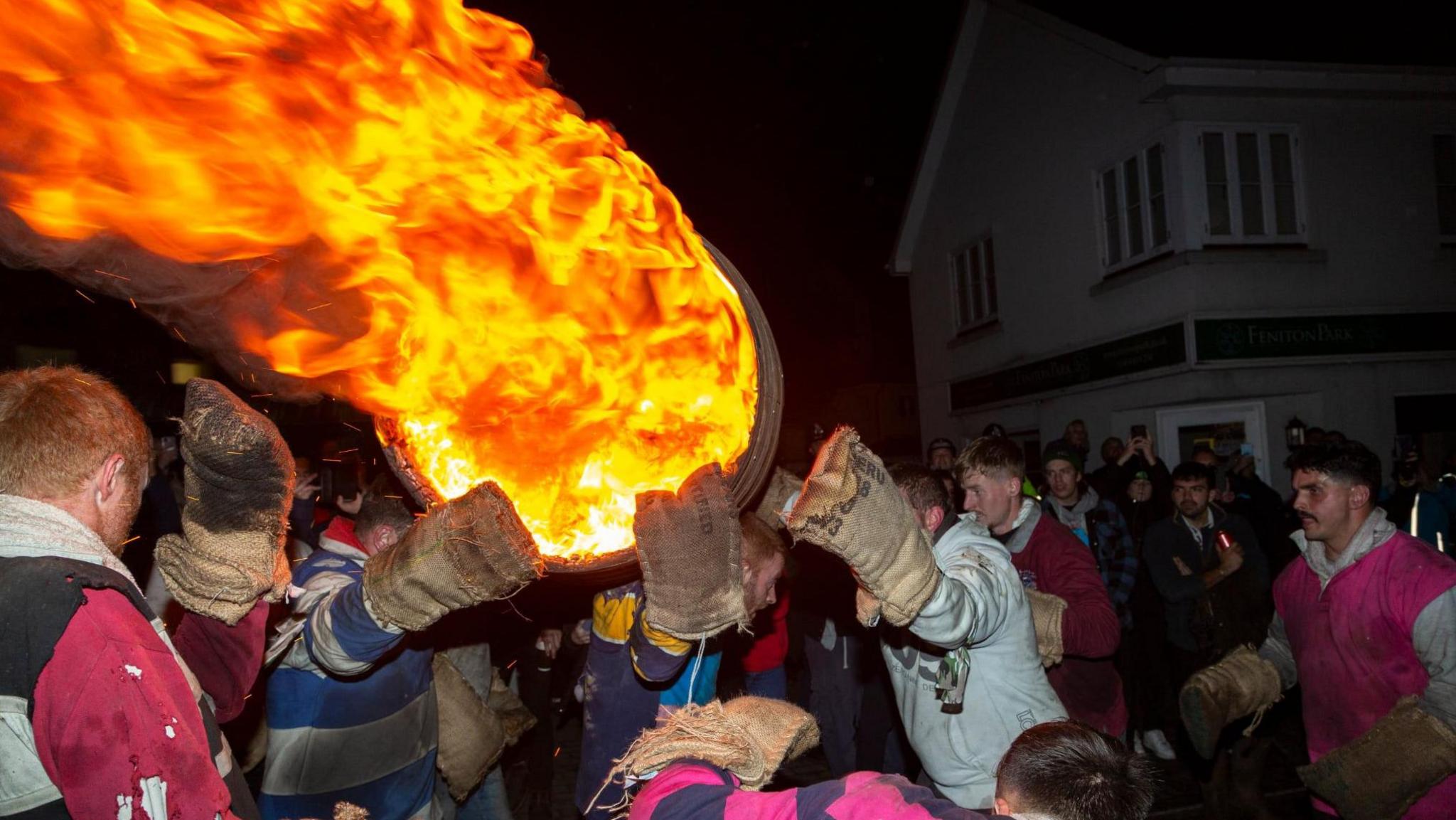 A person carrying a flaming barrel on their shoulders as spectators watch