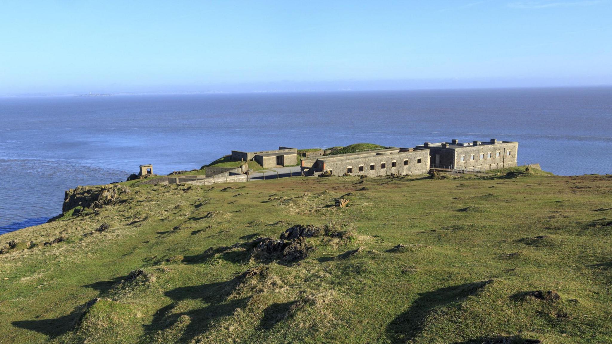 A landscape of grassy, rocky cliffs with the sea in the background. By the edge of the cliff there are a couple buildings - the Palmerston Fort