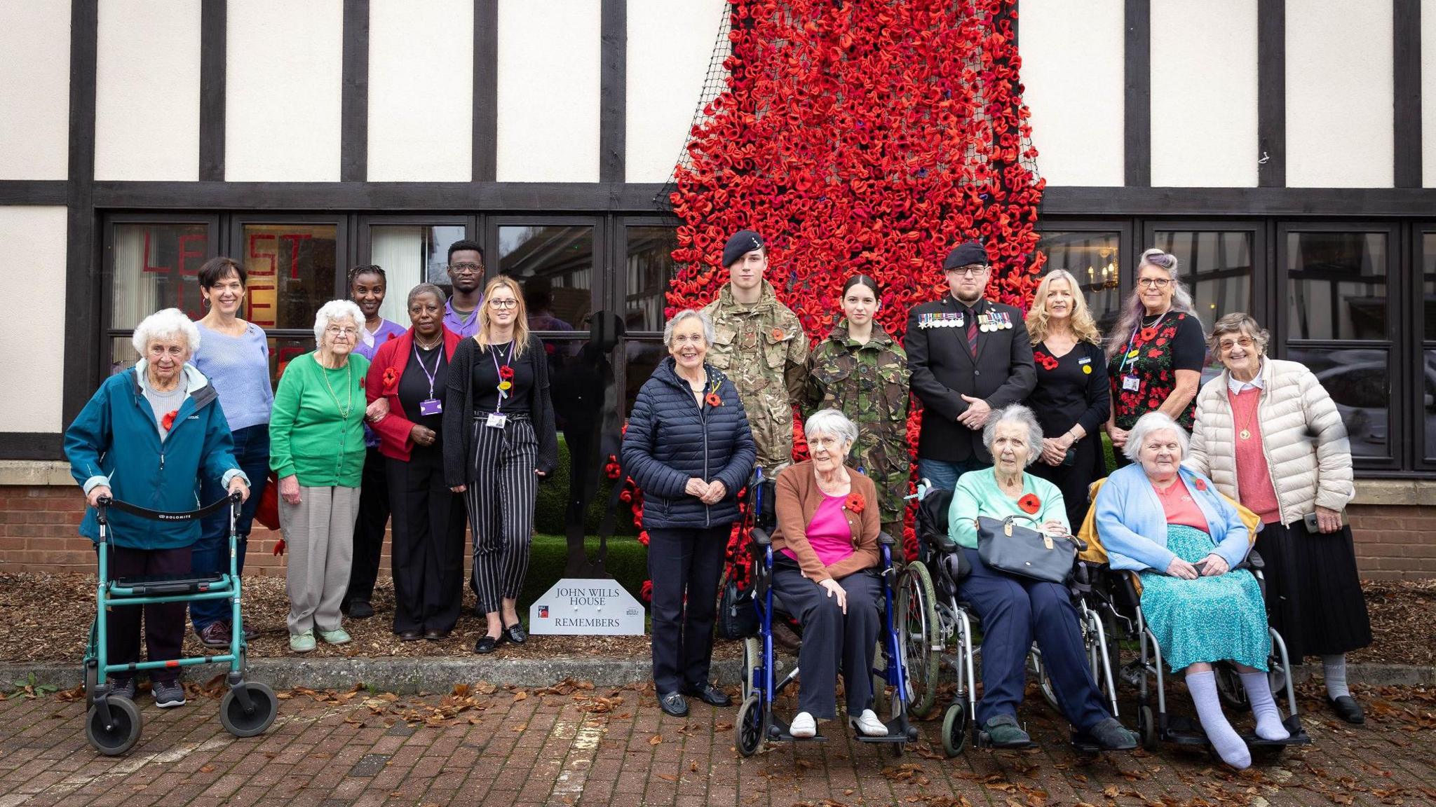 A group of care home residents sit in front of a building under a giant cascade of red crocheted poppies. Some of the people are in wheelchairs and there are representatives of the armed forces standing among them