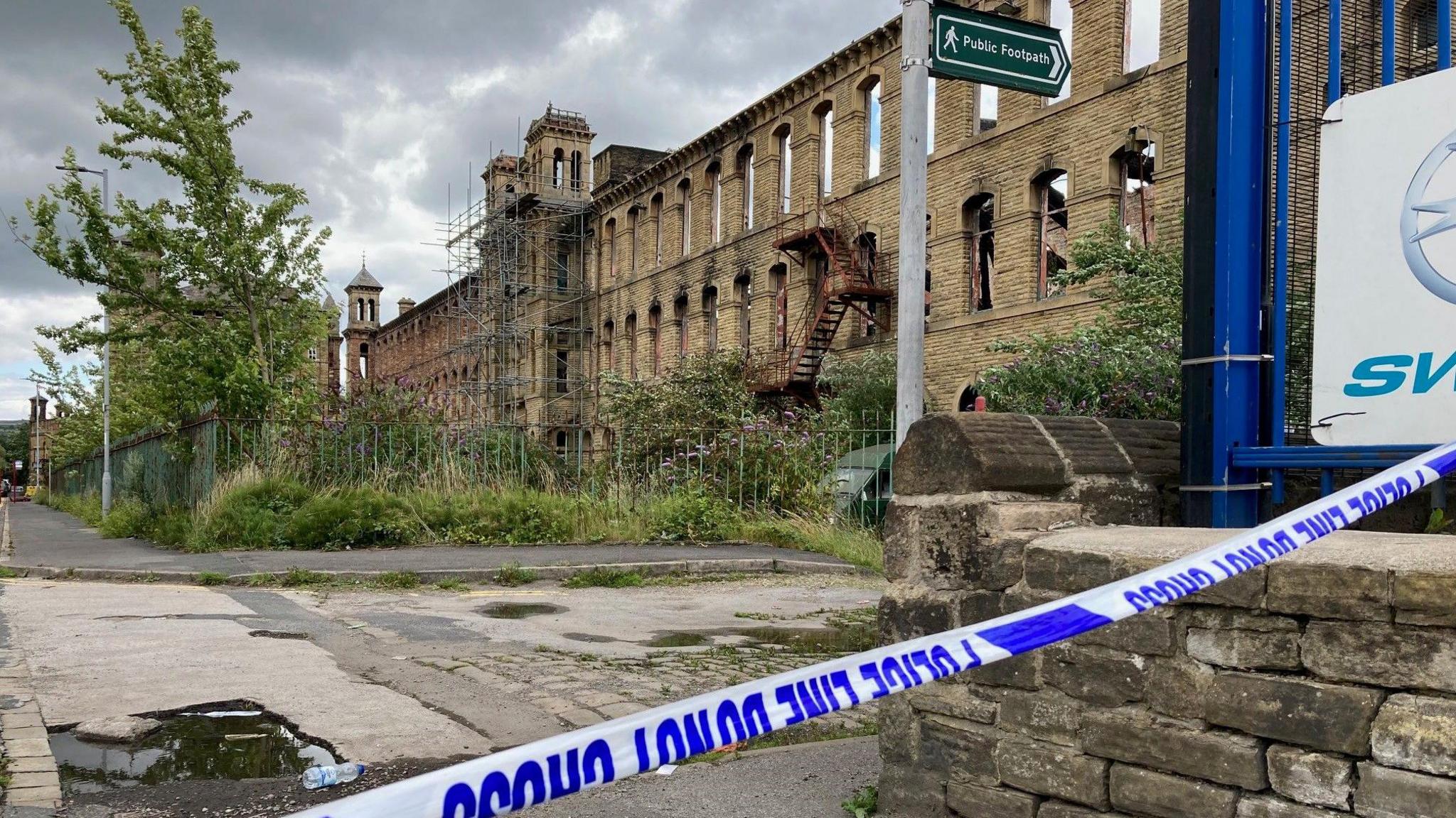 Blue and white police tape with the shell of a burnt-out stone three-storey mill building in the background.