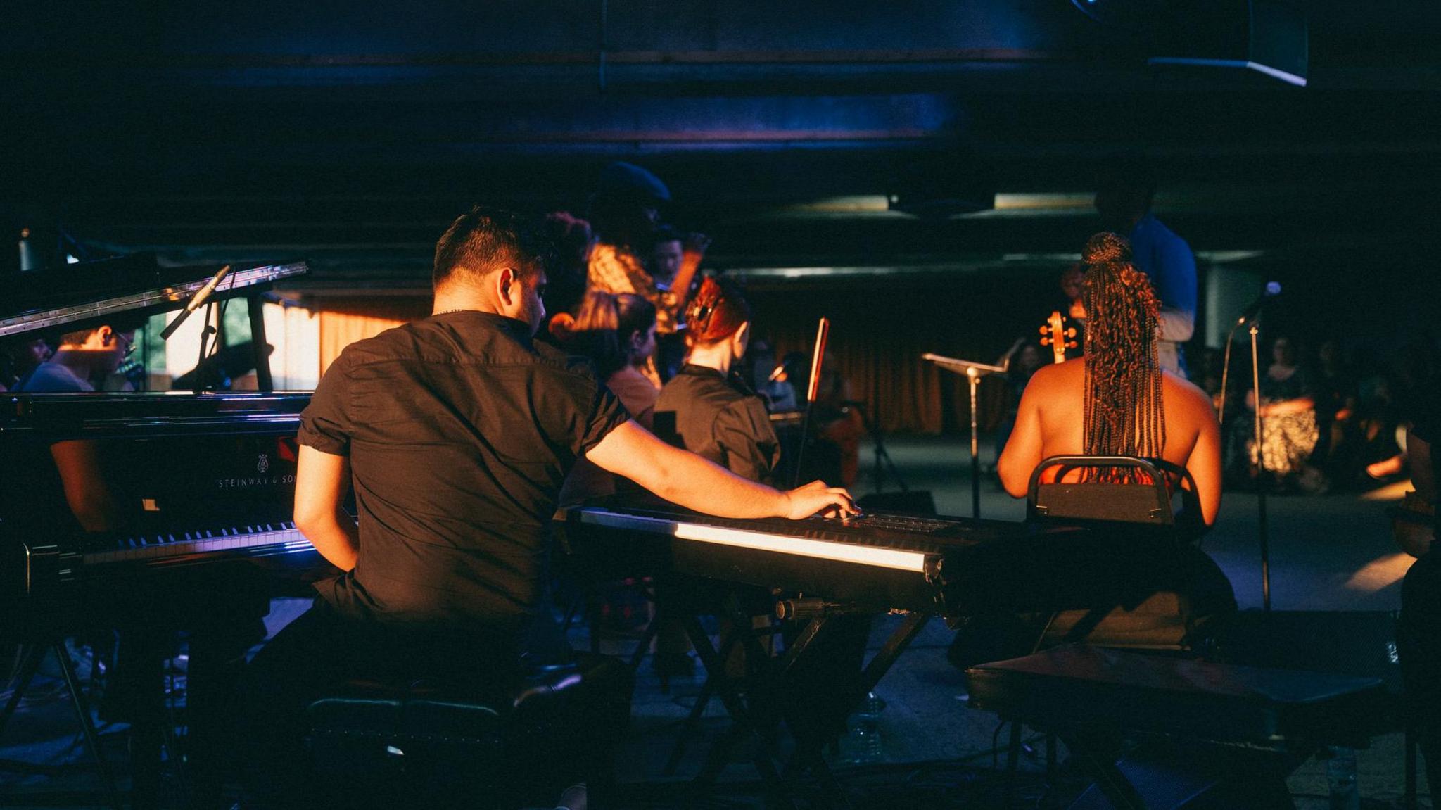 Musicians are facing away from the camera in a dark room. There is a man playing the keyboard in the foreground of the image