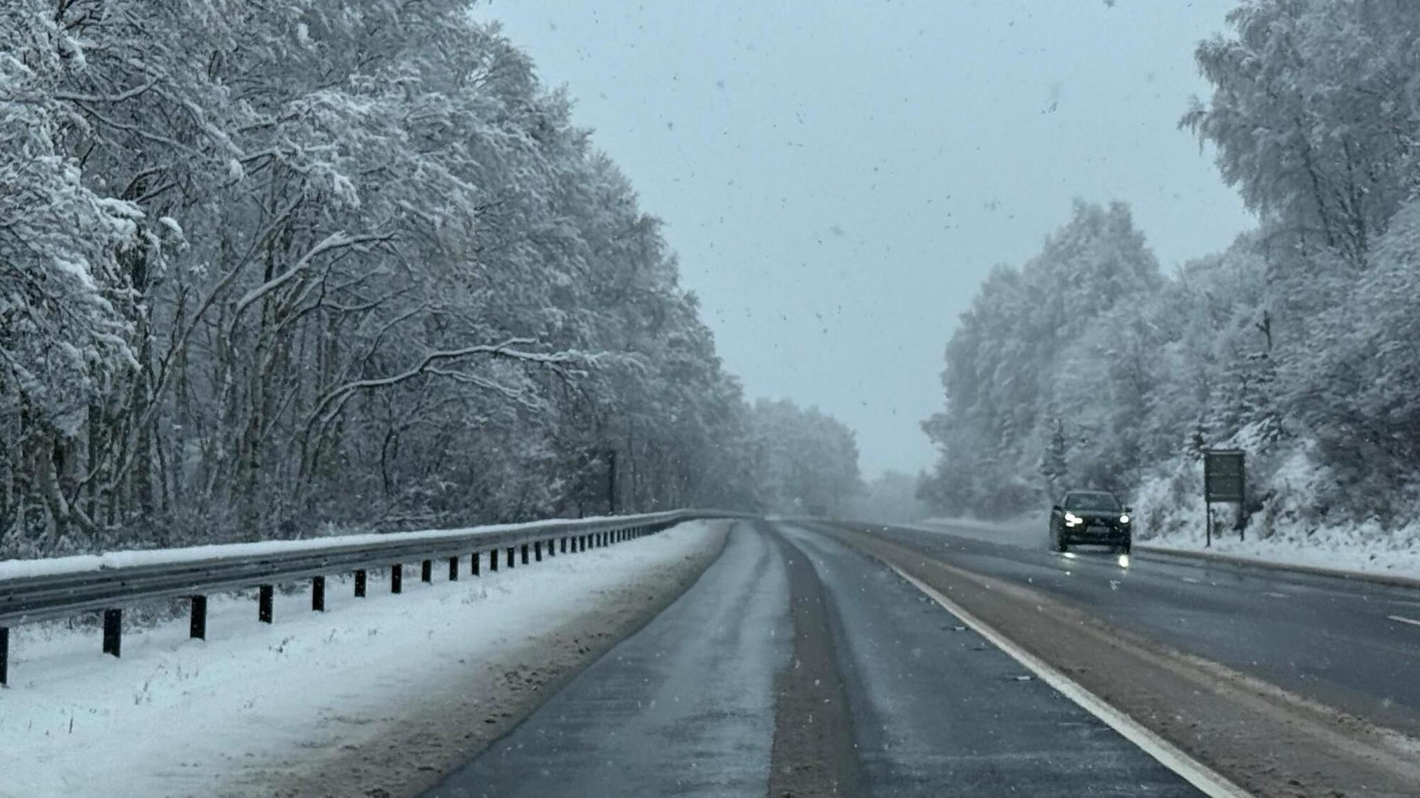 A damp road with evidence of grit and snow pushed to the side. Snow covered trees stand tall on each side and one car approaches with its headlights on in the distance. It is very cloudy and grey