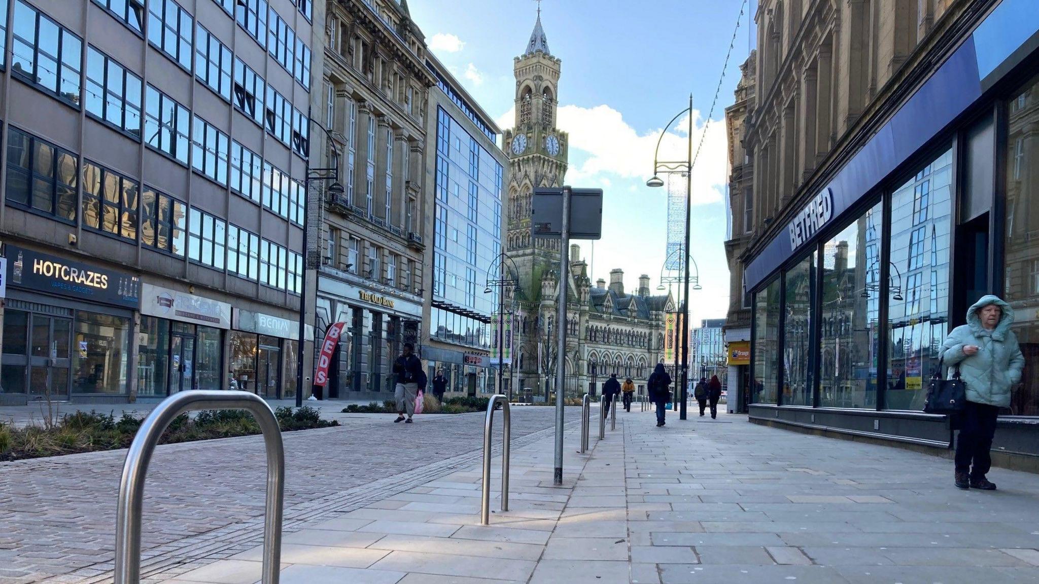 Metal bike security hoops sticking up from a pavement in a long pedestrianised street leading to an ornate 19th century city hall tower in the distance.  