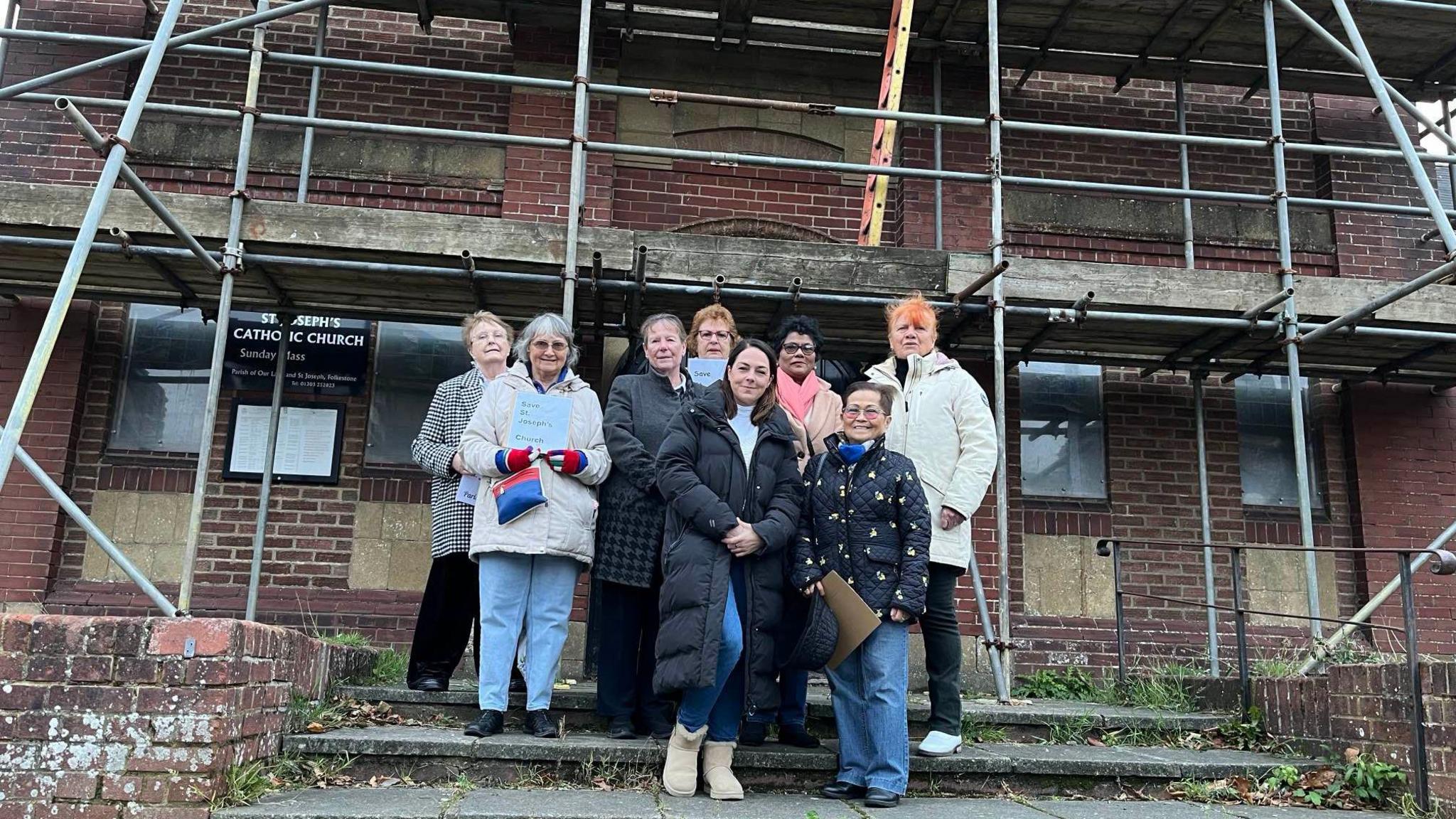 Campaigners stand on the steps at the front of St Joseph's Catholic Church which is made of red brick and covered in scaffolding