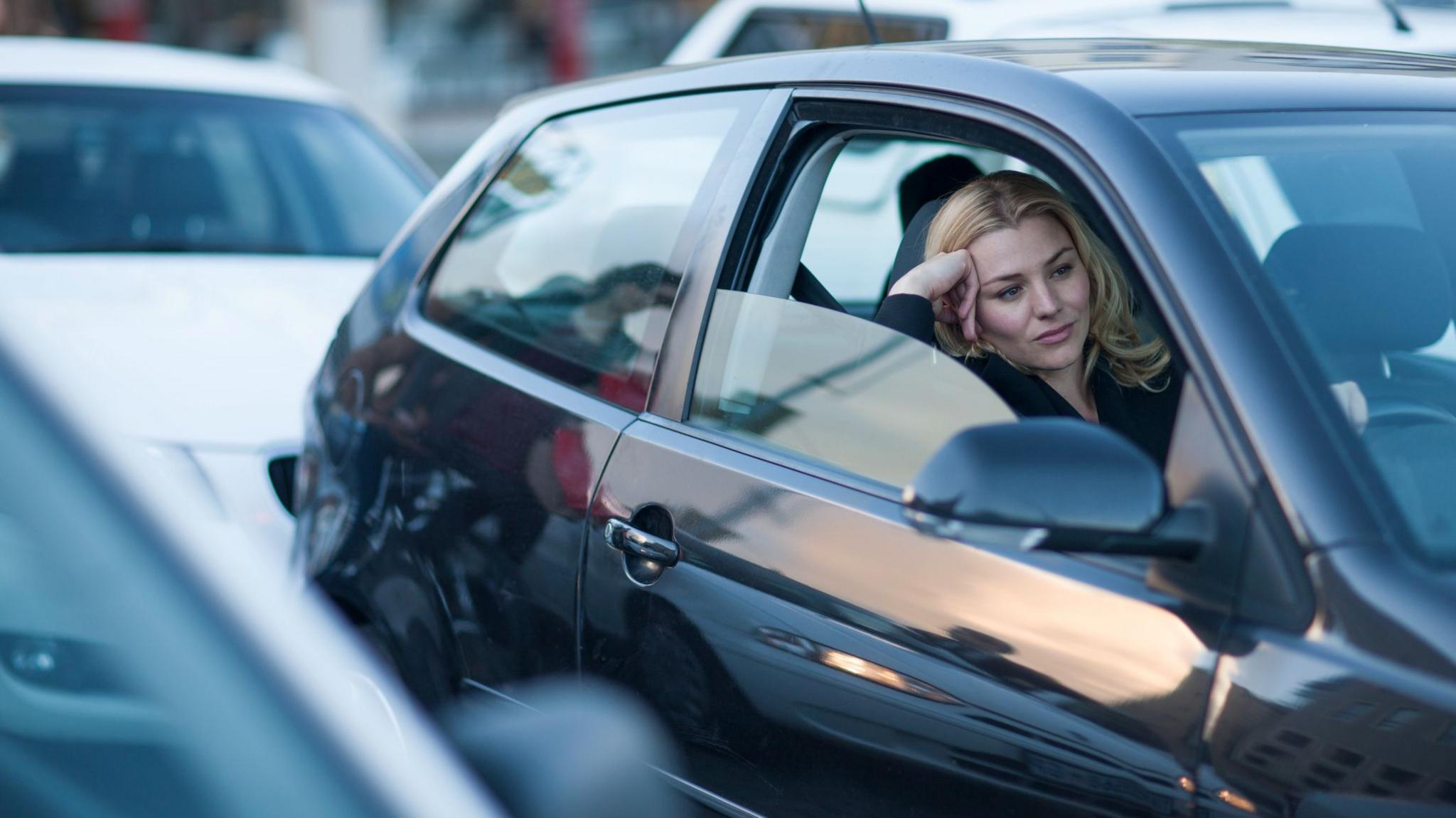 A woman with shoulder length blonde hair sits in her black car in the middle of traffic. She has her window halfway down and is leaning her head on her hand. She looks bored and frustrated.