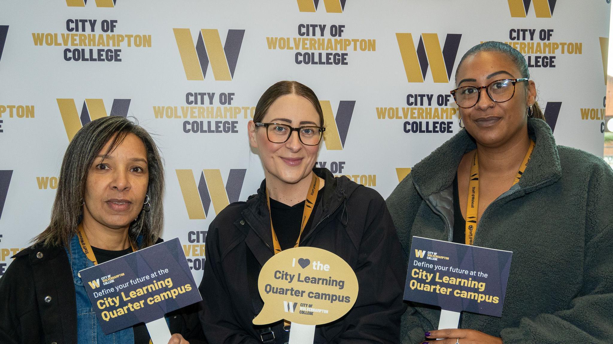Three women smile at the camera and stand in front of a white background with City of Wolverhampton College printed several times on it. They each hold a sign with a slogan about the City Learning Quarter campus on it.