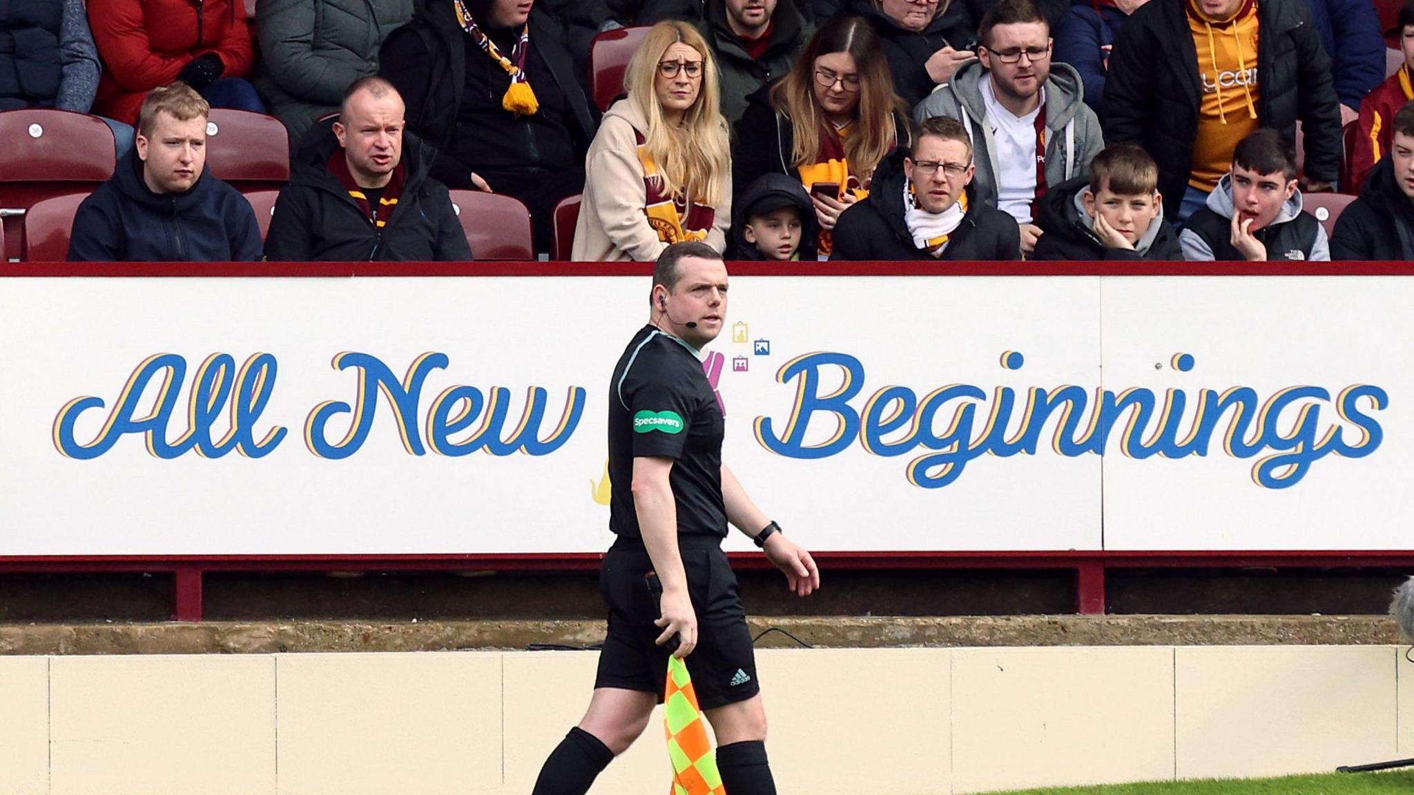 Conservative MSP Douglas Ross (a man with short dark hair) pictured working as a football linesman at what appears to be a Motherwell match. He is wearing all black football kit and holding a yellow and orange flag, and is in front of an advertising board reading "all new beginnings".