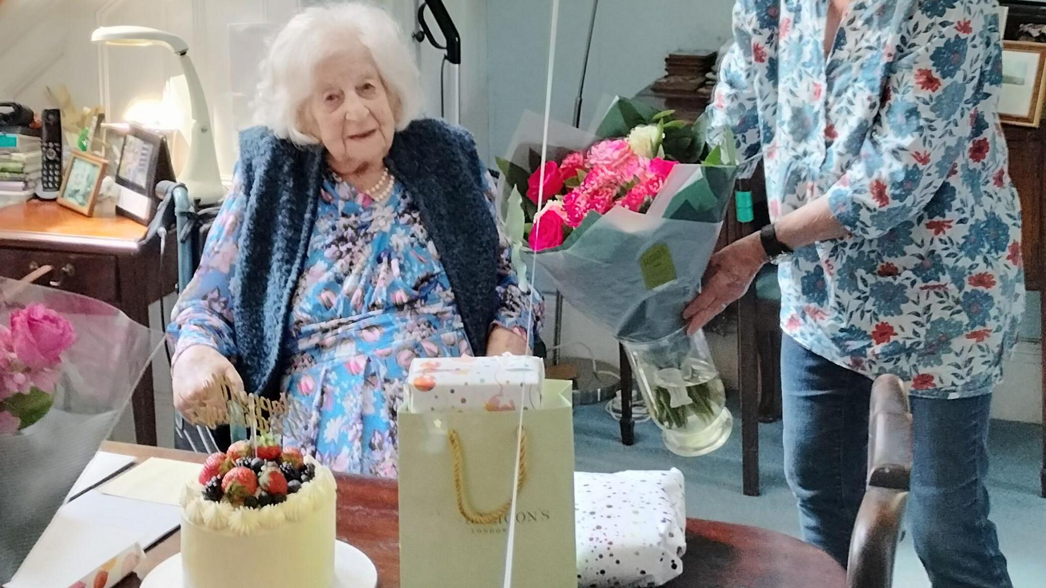 Anne Baker sits in a wheelchair and is wearing a blue dress and pearls. She isbeing presented with bouquets of flowers, a cake and gifts