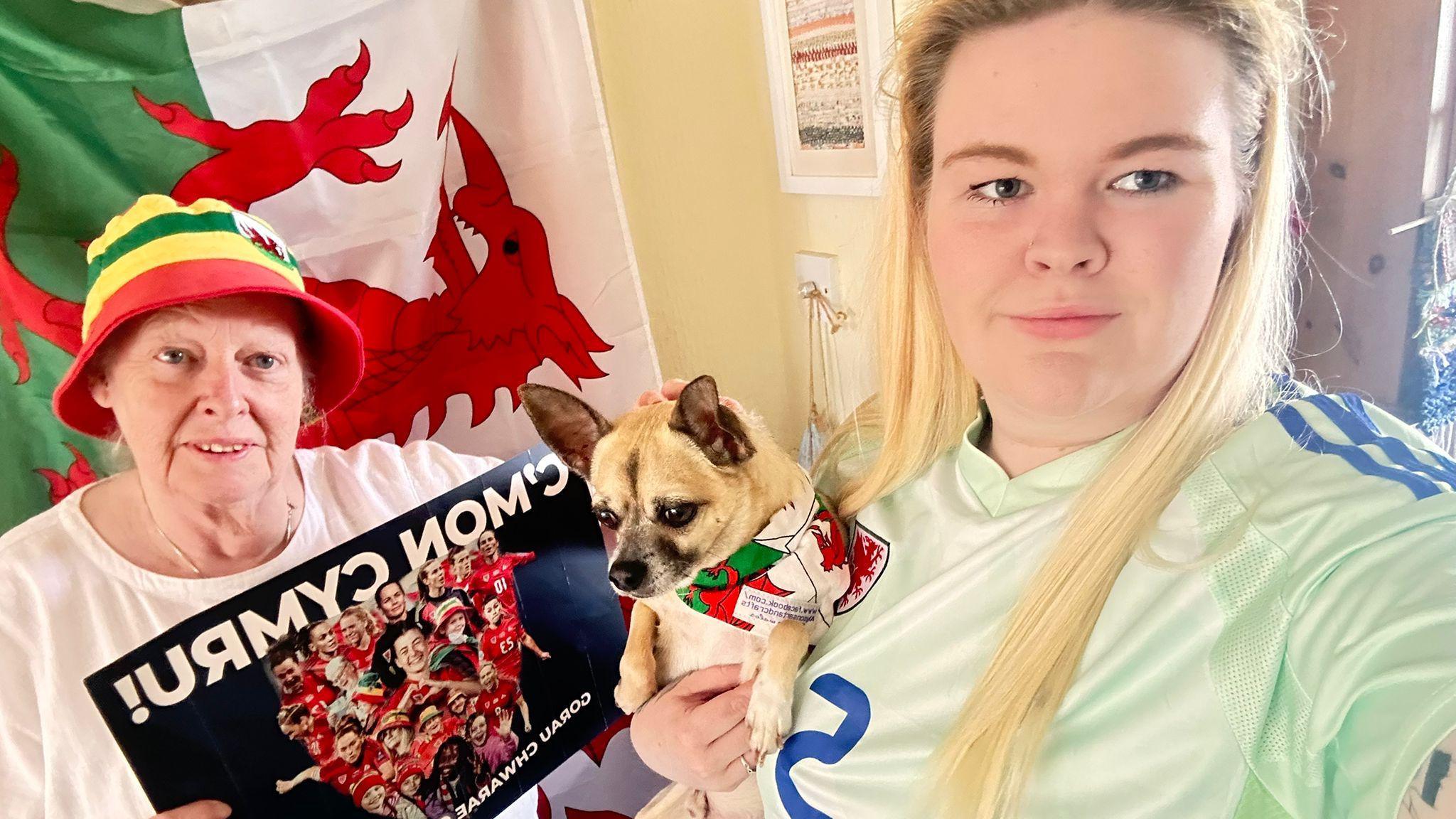 A selfie of Alyson Jane and Hannah Jones and Chester, the dog. Alyson is holding a picture of the team with the words 'C'mon Cymru'. They are standing in front of the Welsh flag.