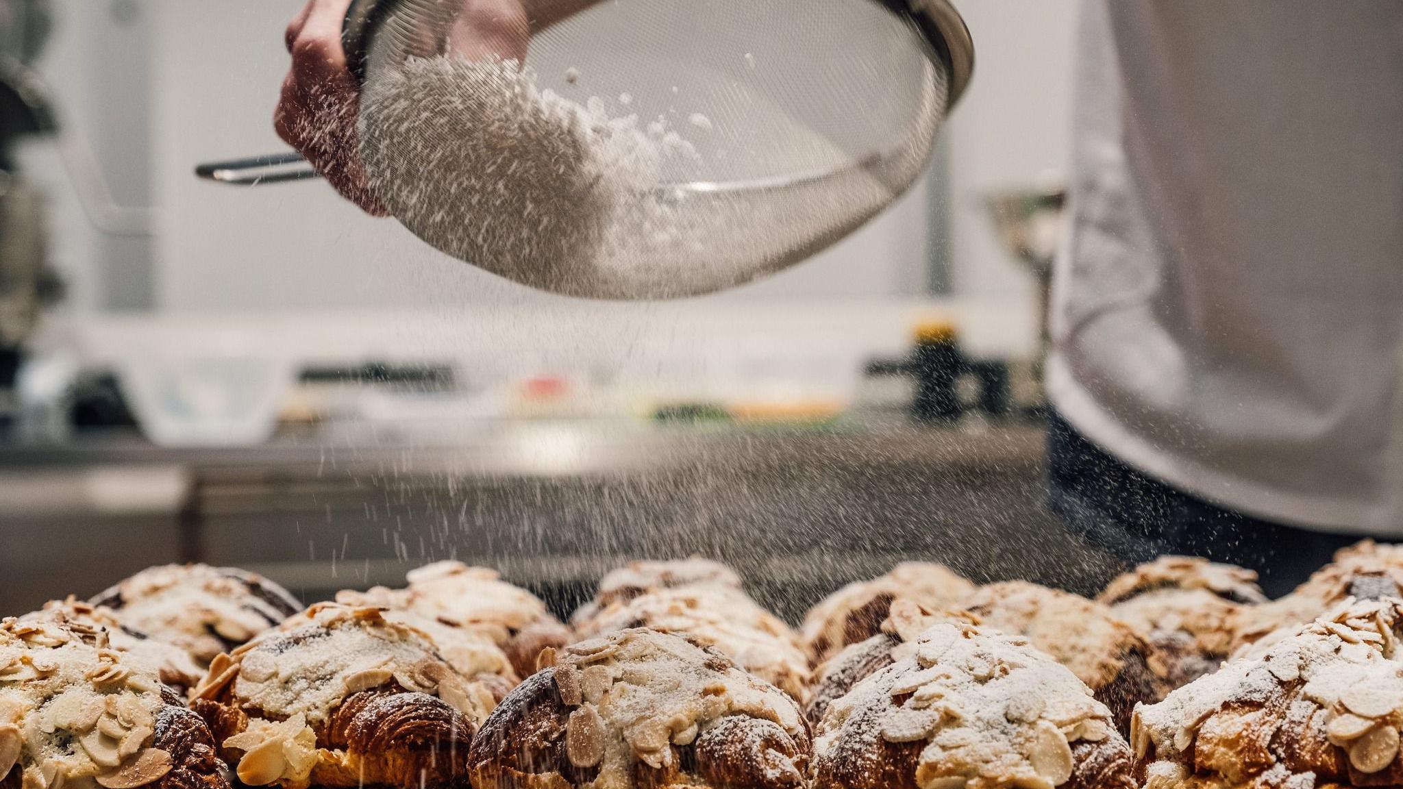 A baker sprinkling powder sugar over almond croissants.