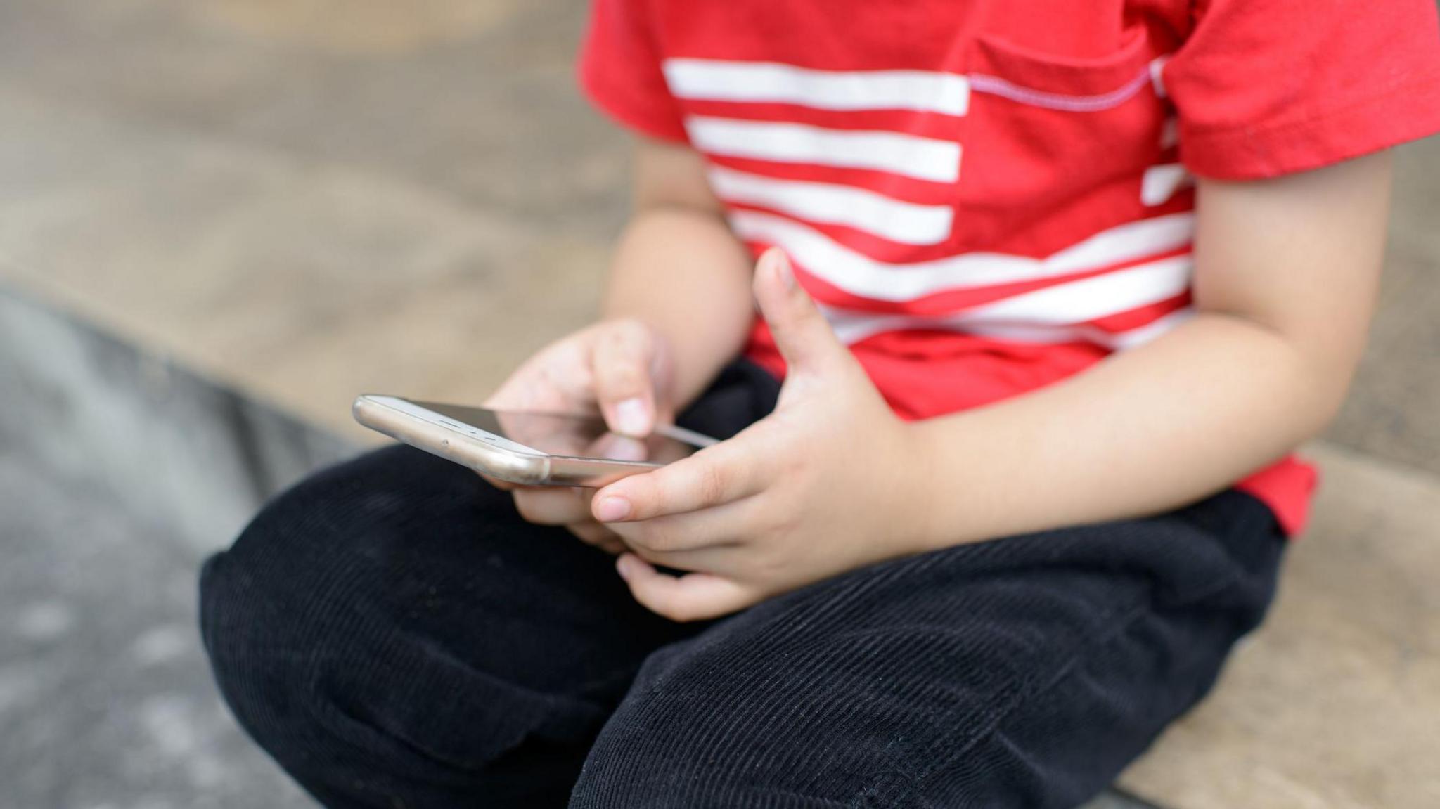 A cropped image of a child holding a smart phone. He wears a red and white striped tshirt and black jeans 