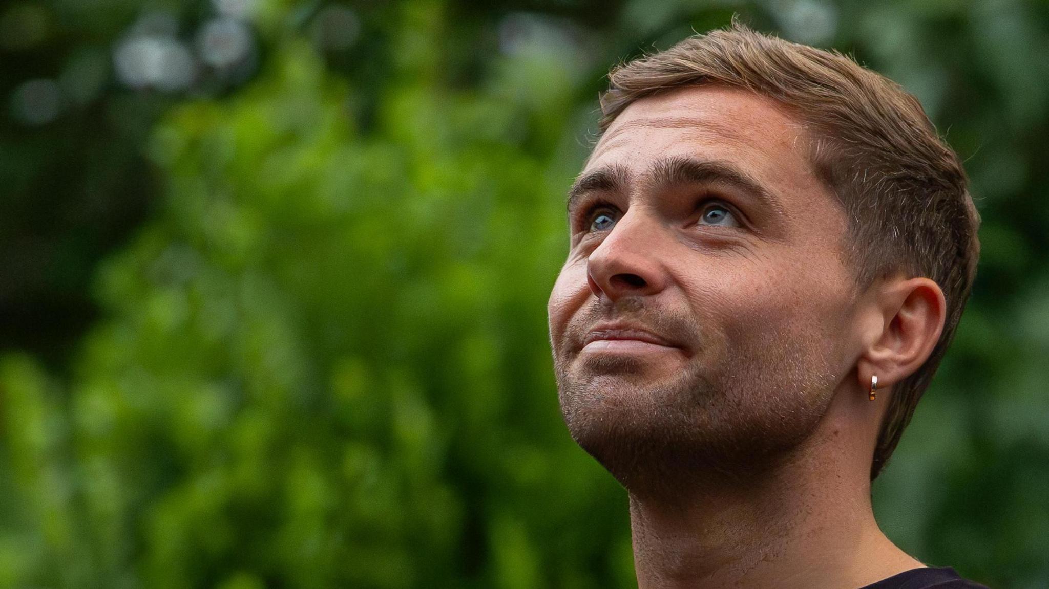 A headshot of Sam King looking upwards while smiling. Green trees can be seen behind him.
