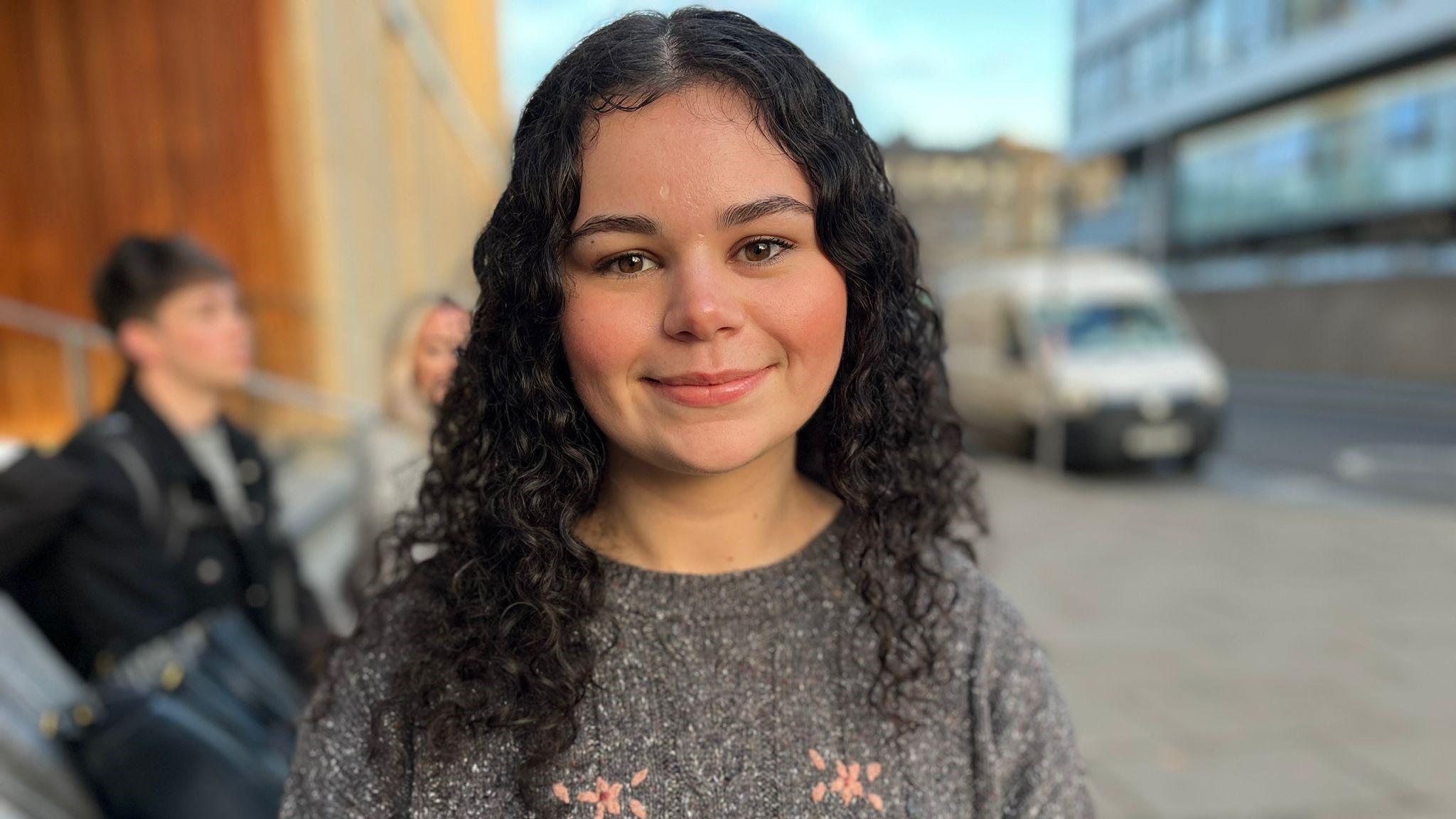 Shanley Breese is wearing a grey jumper with pink flowers on it. She has brown curly hair and is smiling at the camera. There are some other students in the background along with a van and a building.