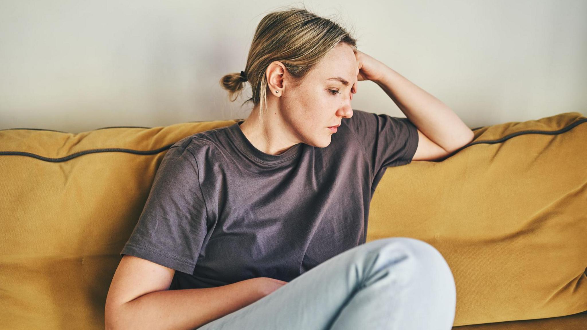 A woman wearing a purple T-shirt and light blue jeans sits on a mustard-yellow sofa and turns her face to the side. She has her head resting on her hand and looks unhappy.