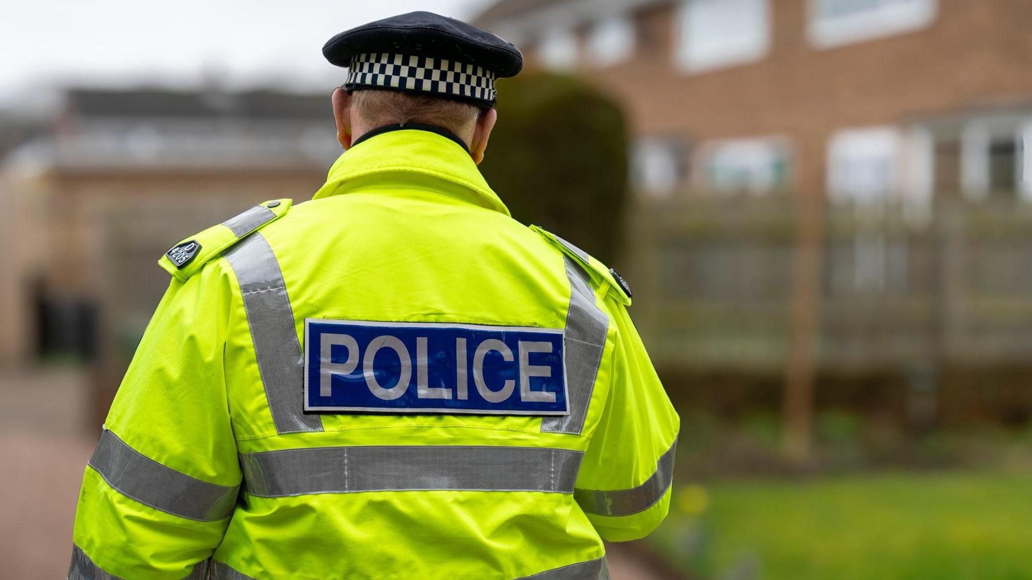 A police officer walks away from the camera, wearing a yellow high viz jacket with silver reflective stripes down the sides and a peaked hat with brown and white check around the rim. The background is blurred but shows a housing estate.