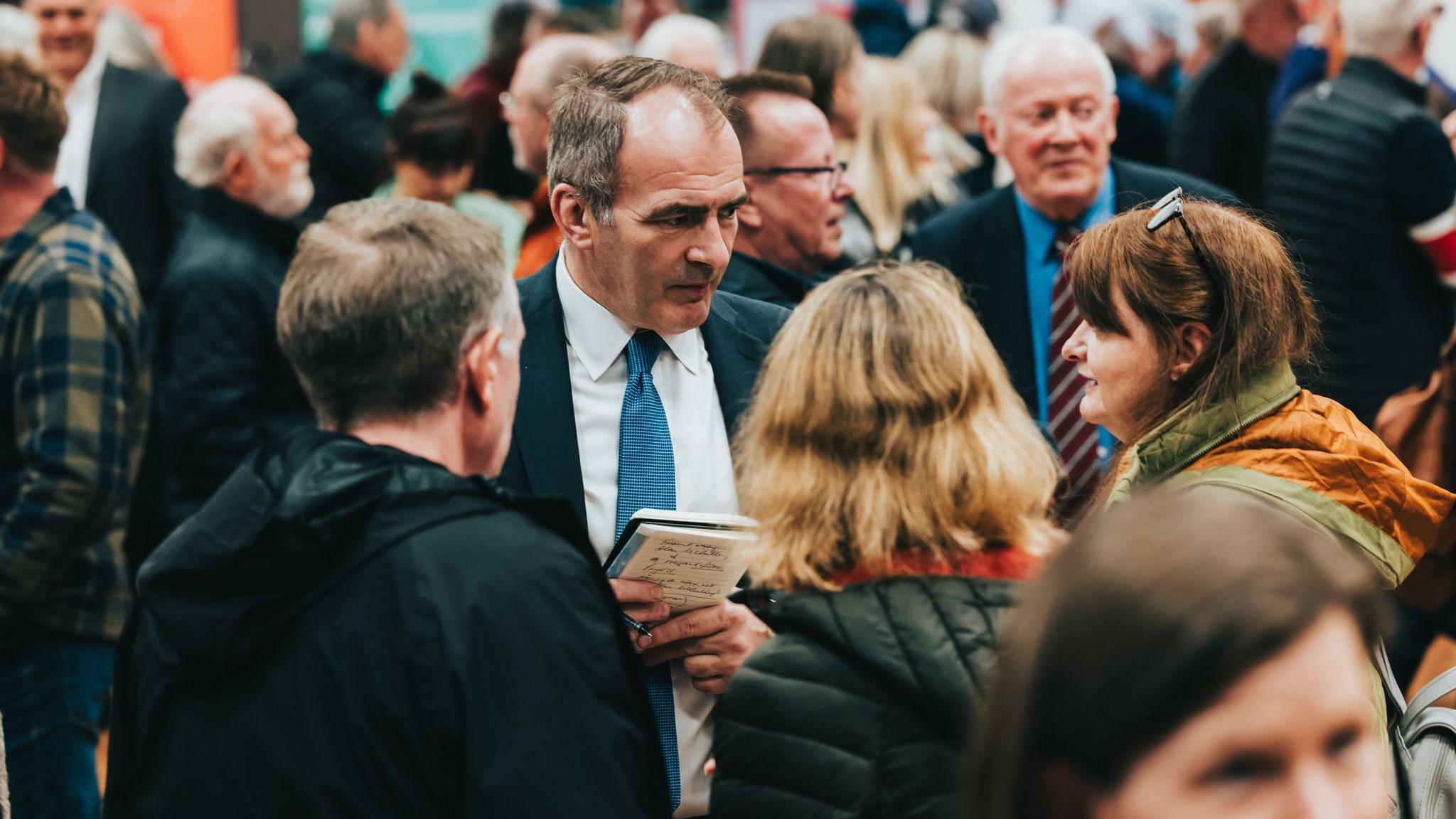 Alfred Cannan stands in the middle of a crowd of people, he talks to a woman wearing a green and yellow coat. He holds a pad and pen in his hands and wears a navy blue suit.