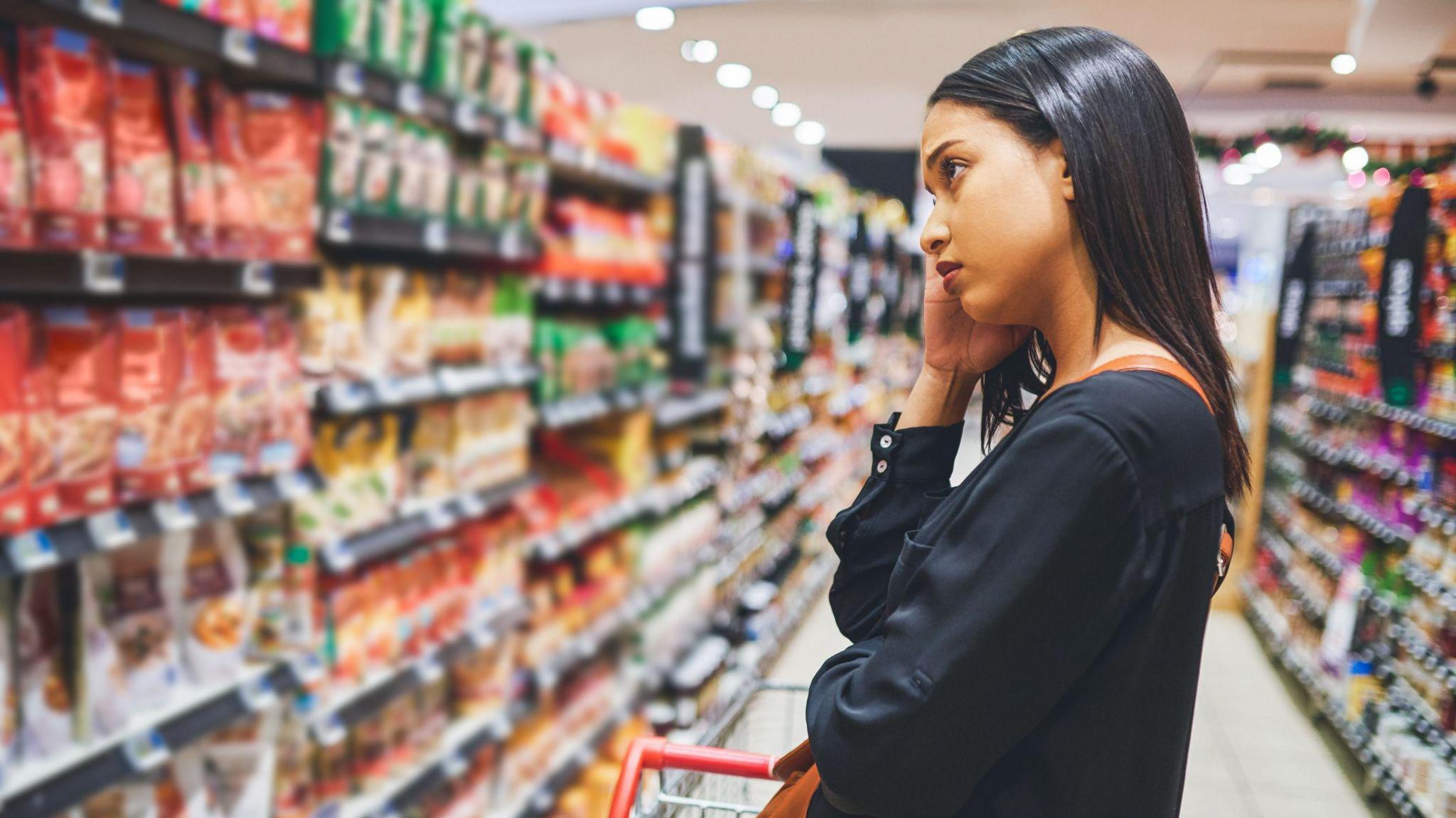 A young woman in a black blouse stands and contemplates a supermarket shelf, with her head leaning on her hand. She has a slightly concerned expression and is carrying a shopping basket.