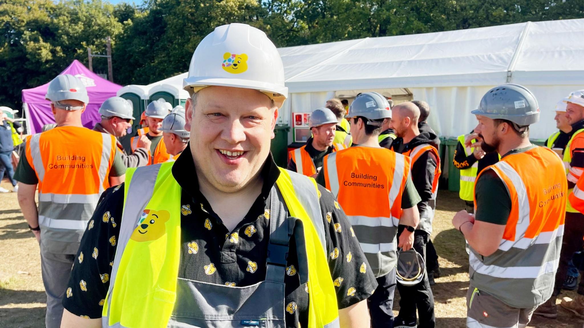 A man in a white hard hat, hi vis vest, black polo shirt and dungarees is standing in front of a dozen other men also dressed in hard hats and hi vis vests. The man's hard hat, hi-vis vest and polo shirt are decorated with imnages of Children in Need mascot, Pudsey, a yellow bear with a spotty eyepatch. Behind all the men is a large white marquee, a smaller purple marquee and tall green trees. It is a sunny day.