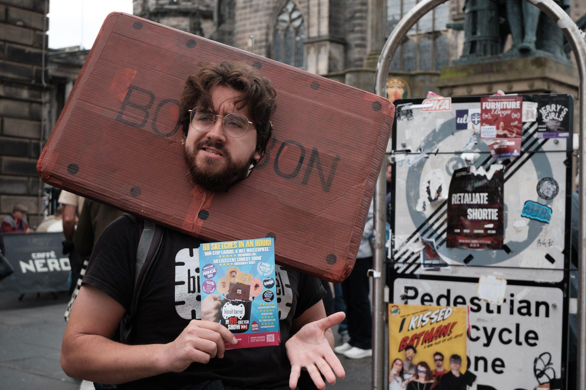 A dark-haired bearded man wears a Bourbon biscuit headpiece as he gives out flyers on the Royal Mile