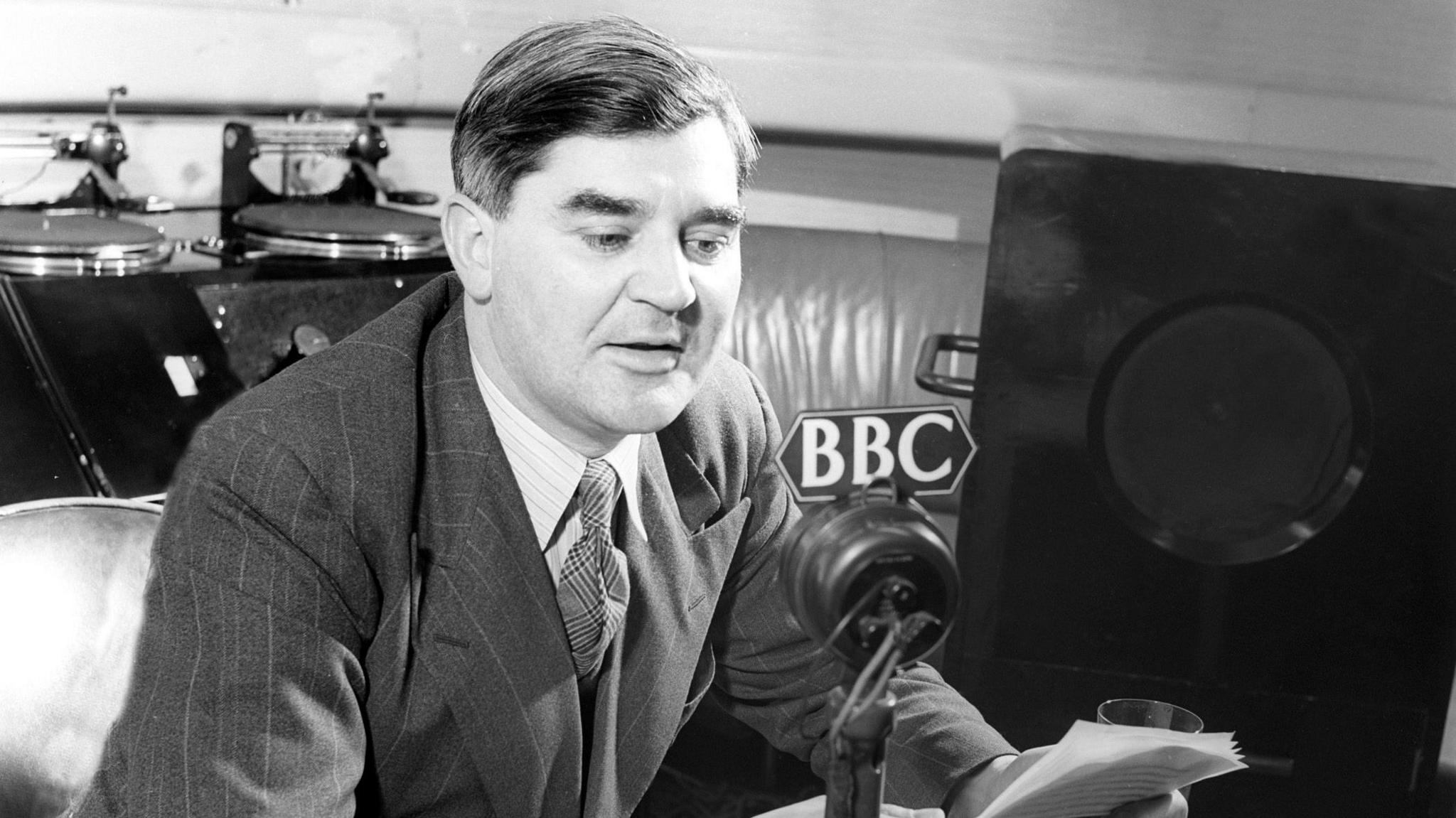 A black and white image of Nye Bevan delivering a broadcast on the radio. The microphone is branded with the BBC logo.