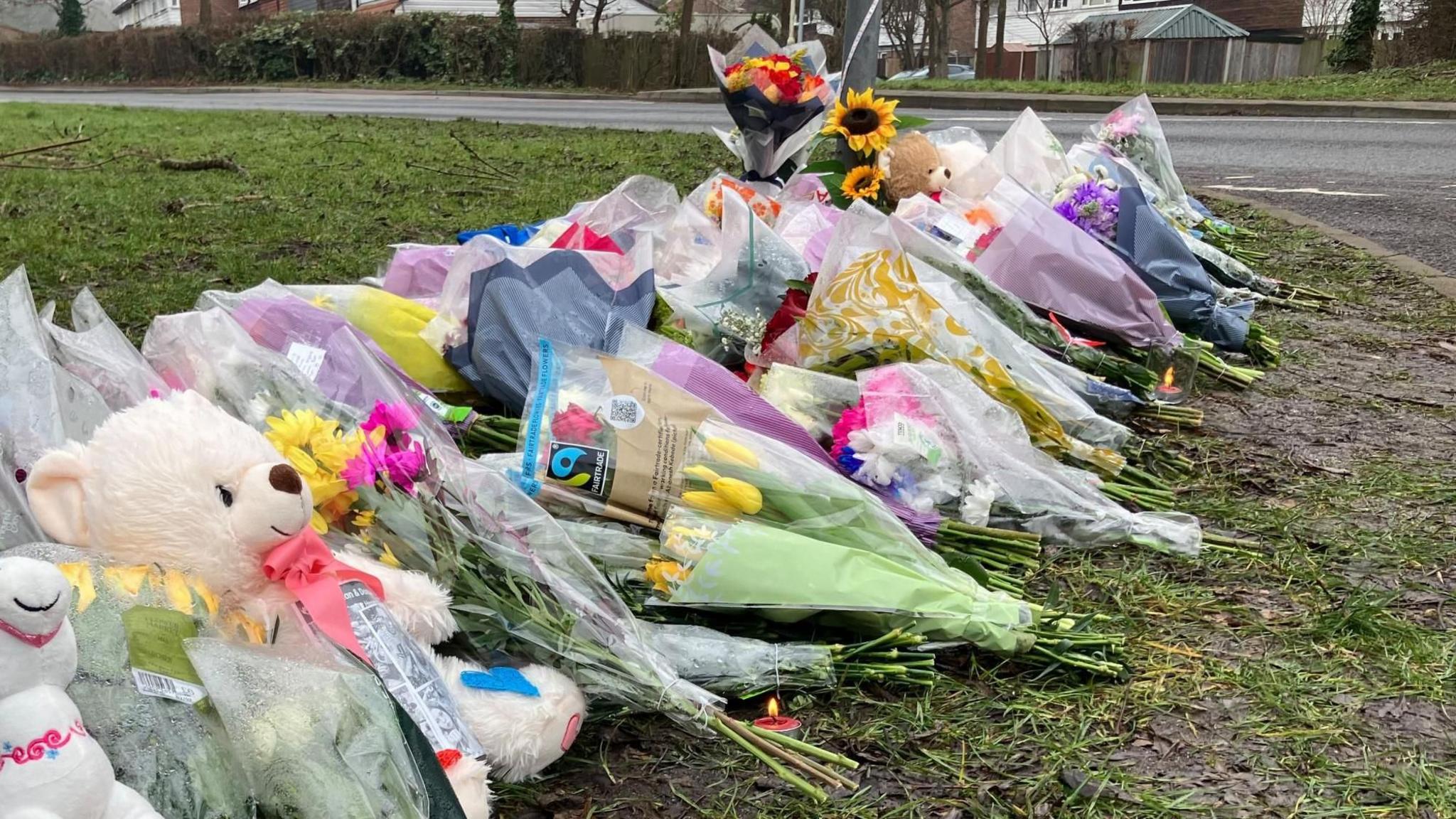 Bunches of flowers are lying on grass at the side of a road. Some have messages written on them