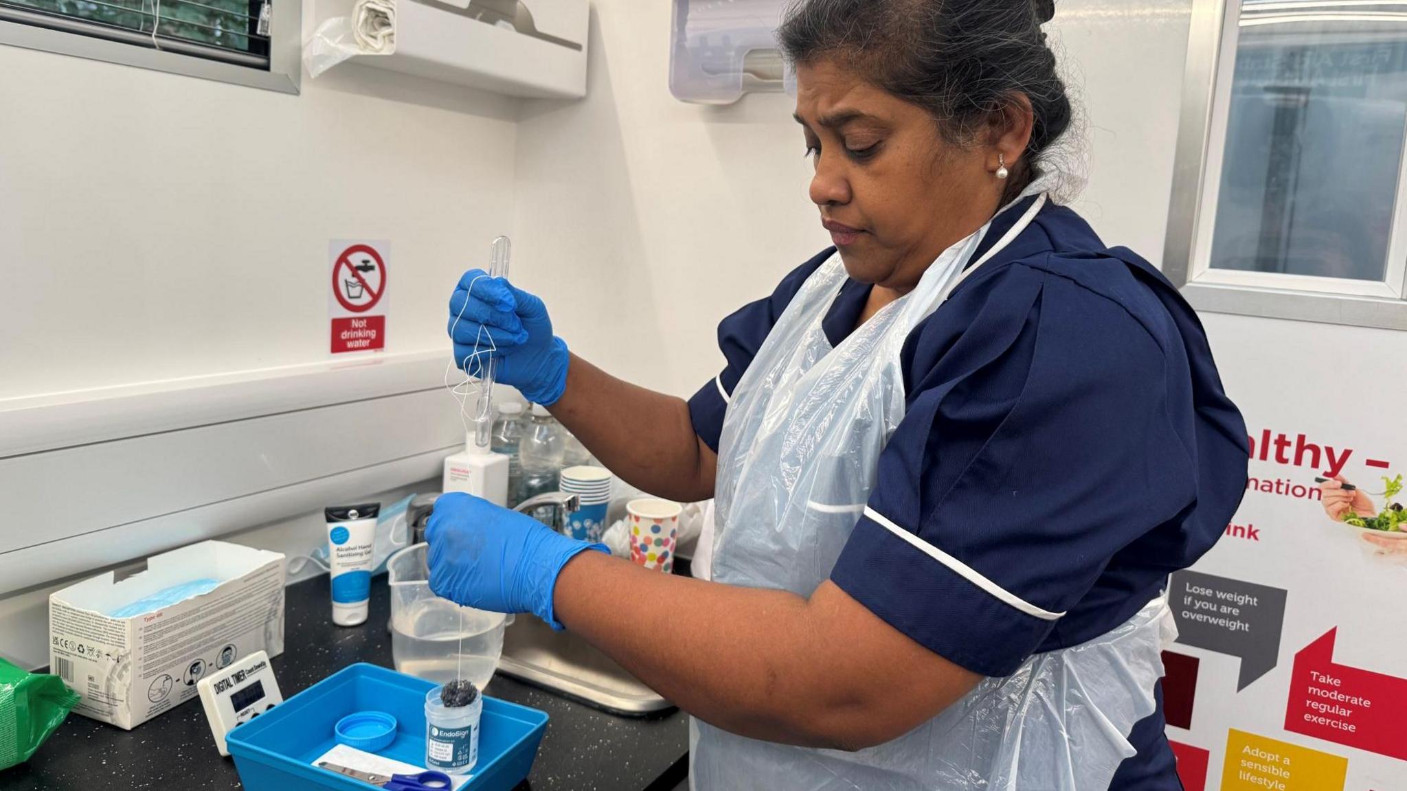 A nurse in a navy uniform with white trim, wearing a disposable apron and blue gloves, prepares a medical test using a sponge with string attached and a blue tray of supplies. The clinical setting includes a digital timer, test kit, and organised equipment.