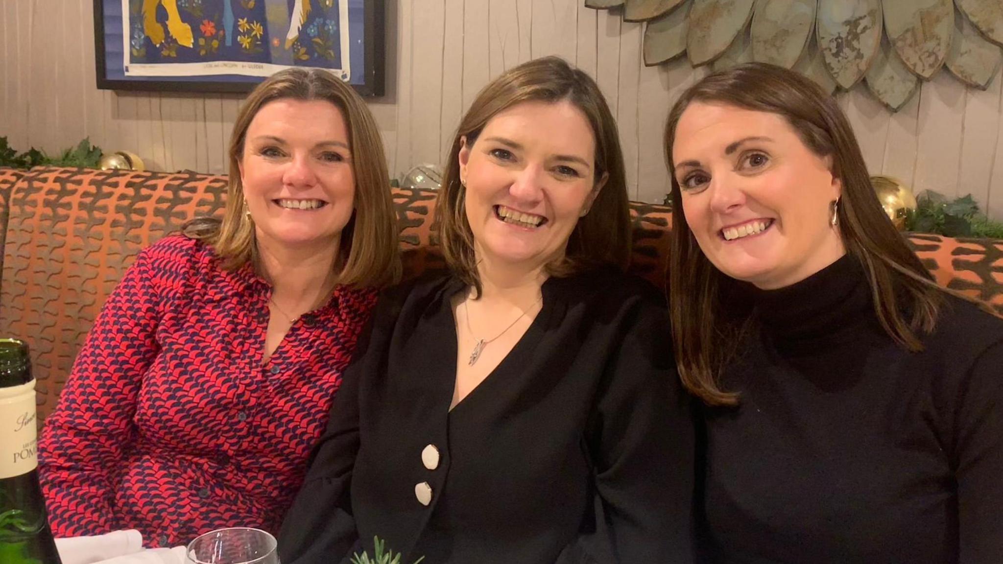 The picture shows three woman all with shoulder length brown hair smiling while sitting on a patterned sofa 