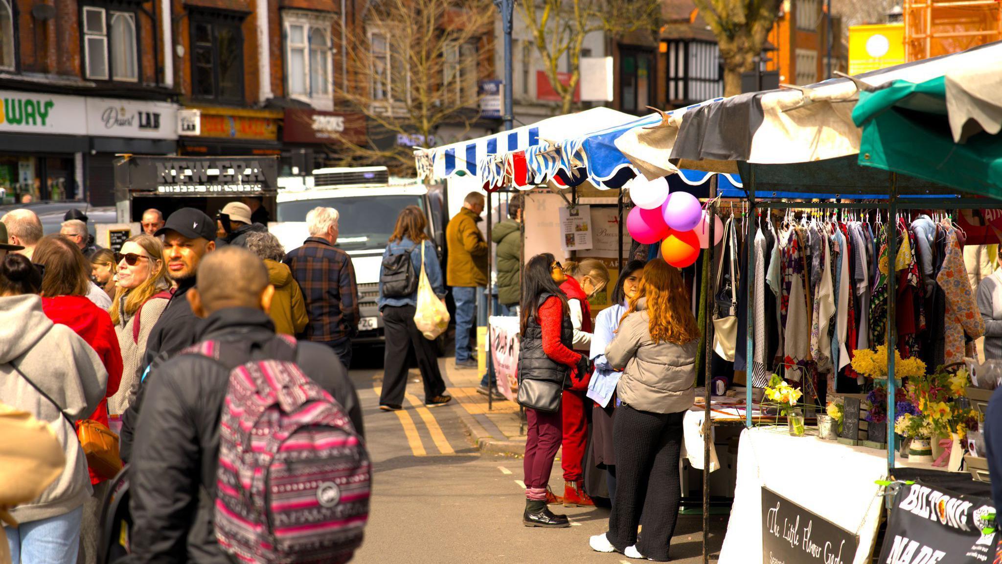 A general view of the market with crowds of people walking around the stalls that line both sides of the street. There are shops in the distance too. 