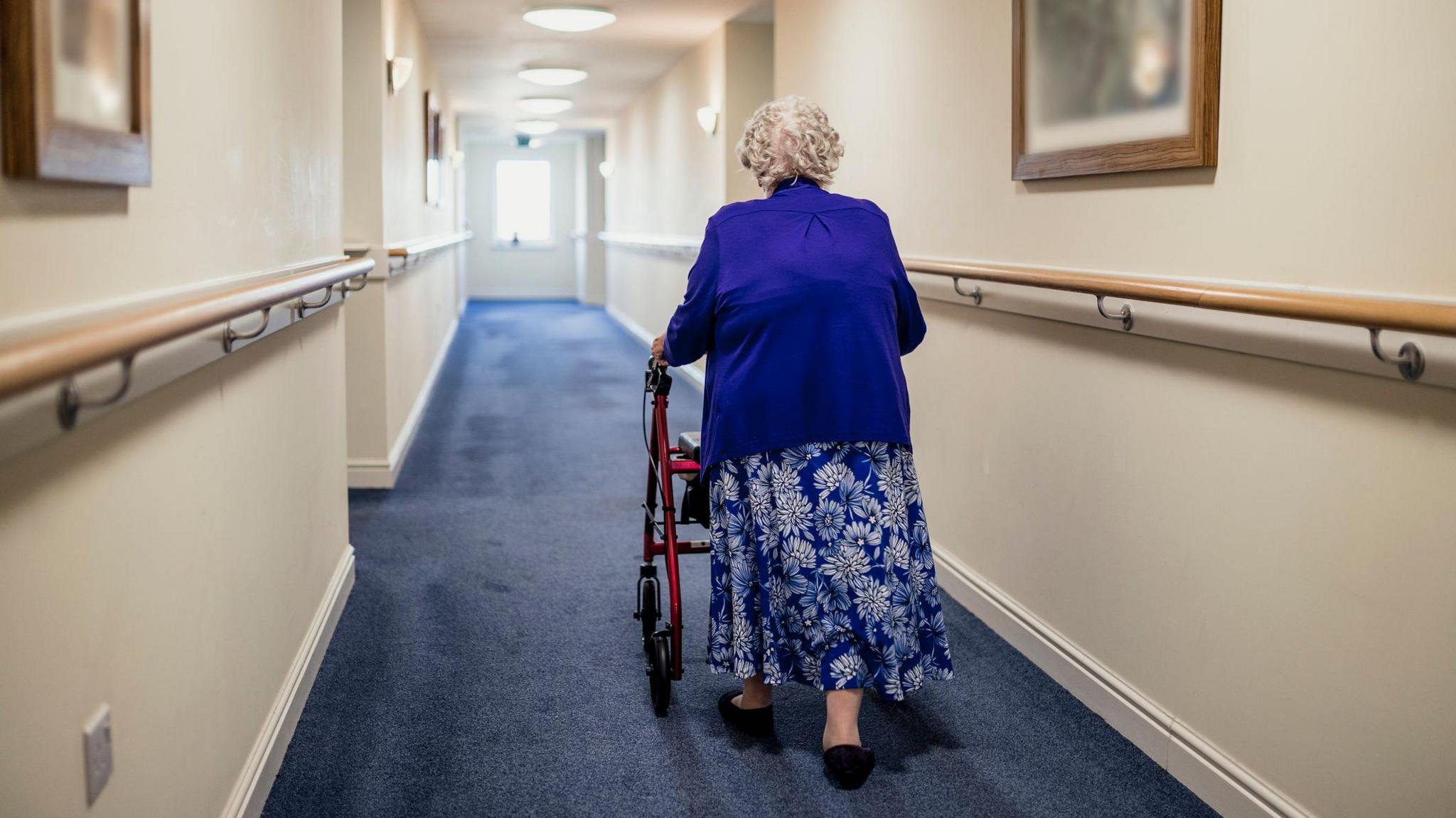 An elderly woman walks down a corridor in a care home with her back to the camera. She is using a walker for support and is wearing a blue cardigan and a long blue flowery skirt. The walls are cream and the carpet is blue.