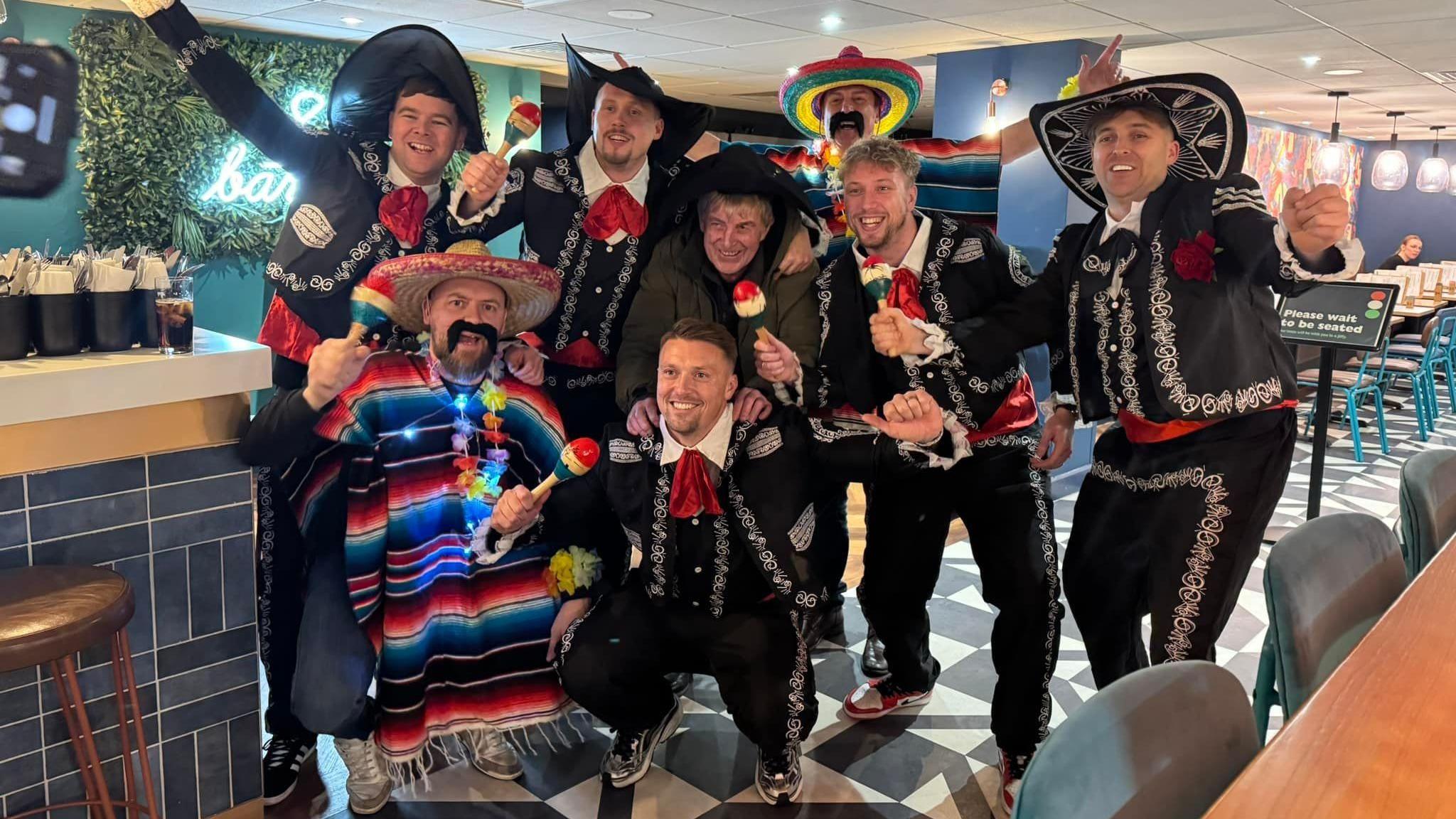 A group of eight men strike celebratory poses inside a venue with customer tables. Seven of them wear Mexican costumes, including embroidered jackets, a cape or sombreros. An older man in the centre is wearing a hat but not the other fancy dress items