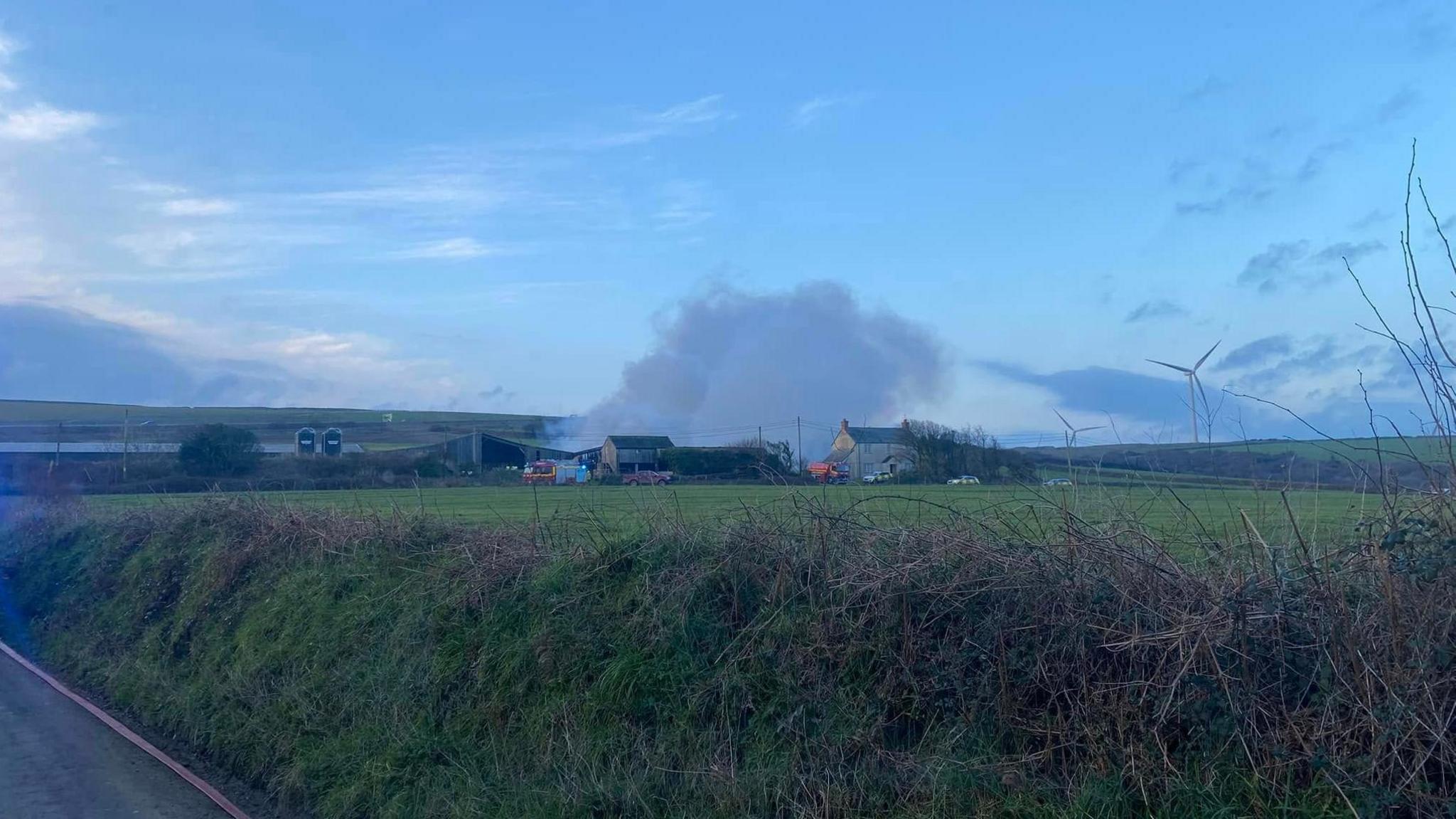 A view fop the farm from distance. A mass of smoke is billowing into the sky. There are two fire engines and three emergency vehicles visible next to the cluster of farm buildings. There is a hedge and a road in the foreground and a field between the person taking the photo and the farm.