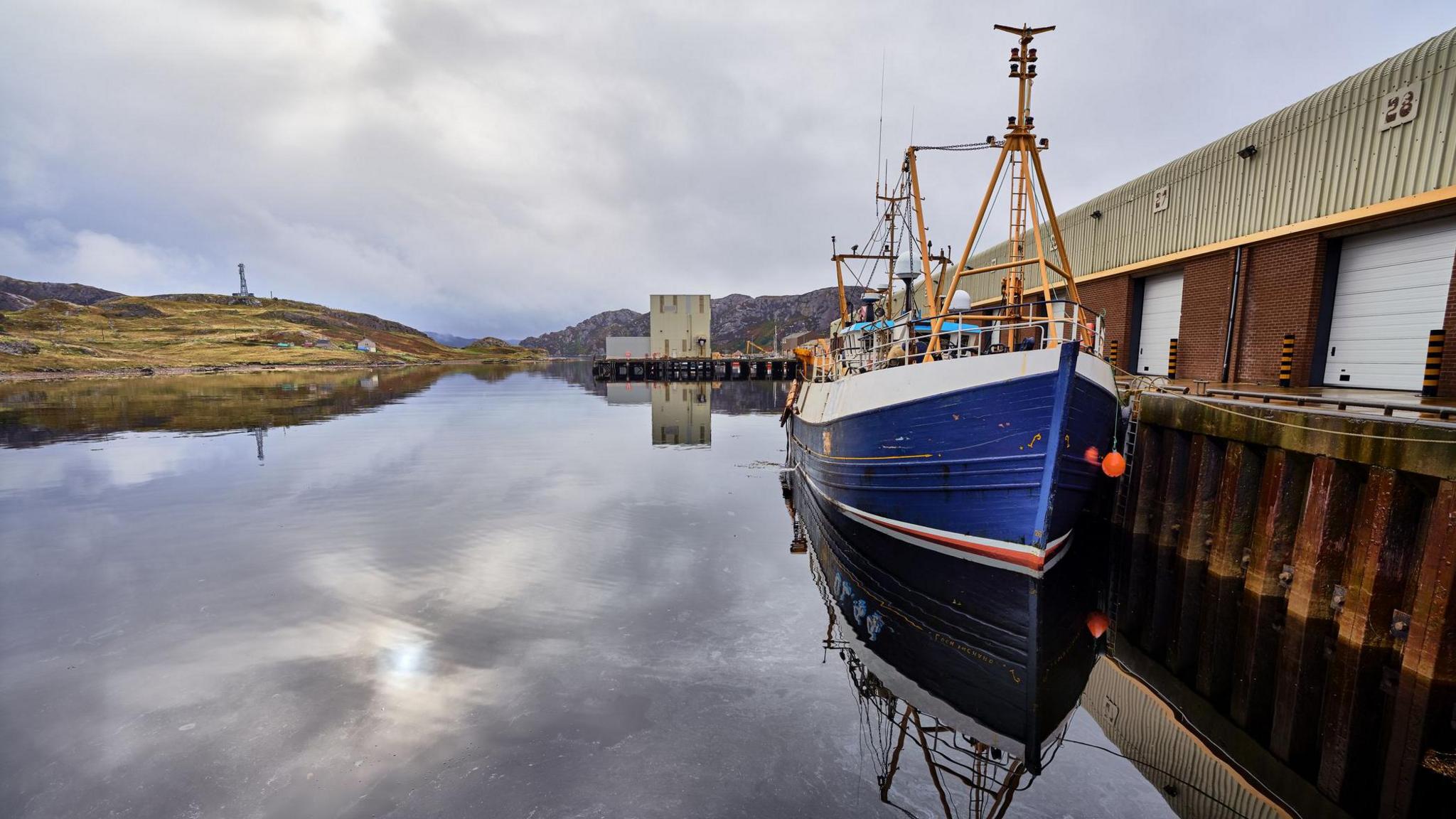 A blue and white hulled fishing boat is tied up in the harbour. The water is flat calm and reflects the cloudy sky above. There is a large harbour building and rocky hills in the distance.