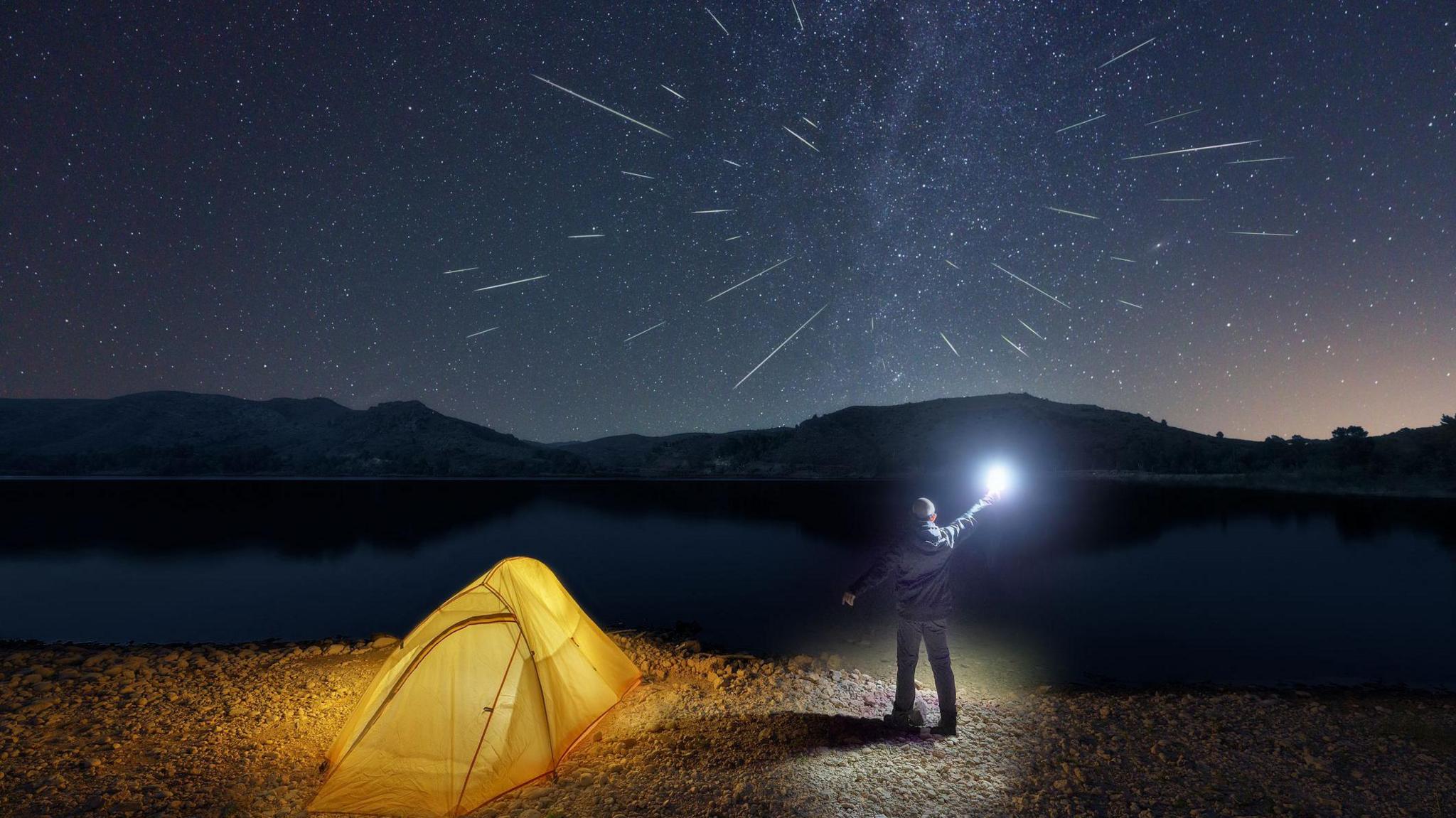 man with a lantern next to his camping tent is watching the Perseid meteor shower