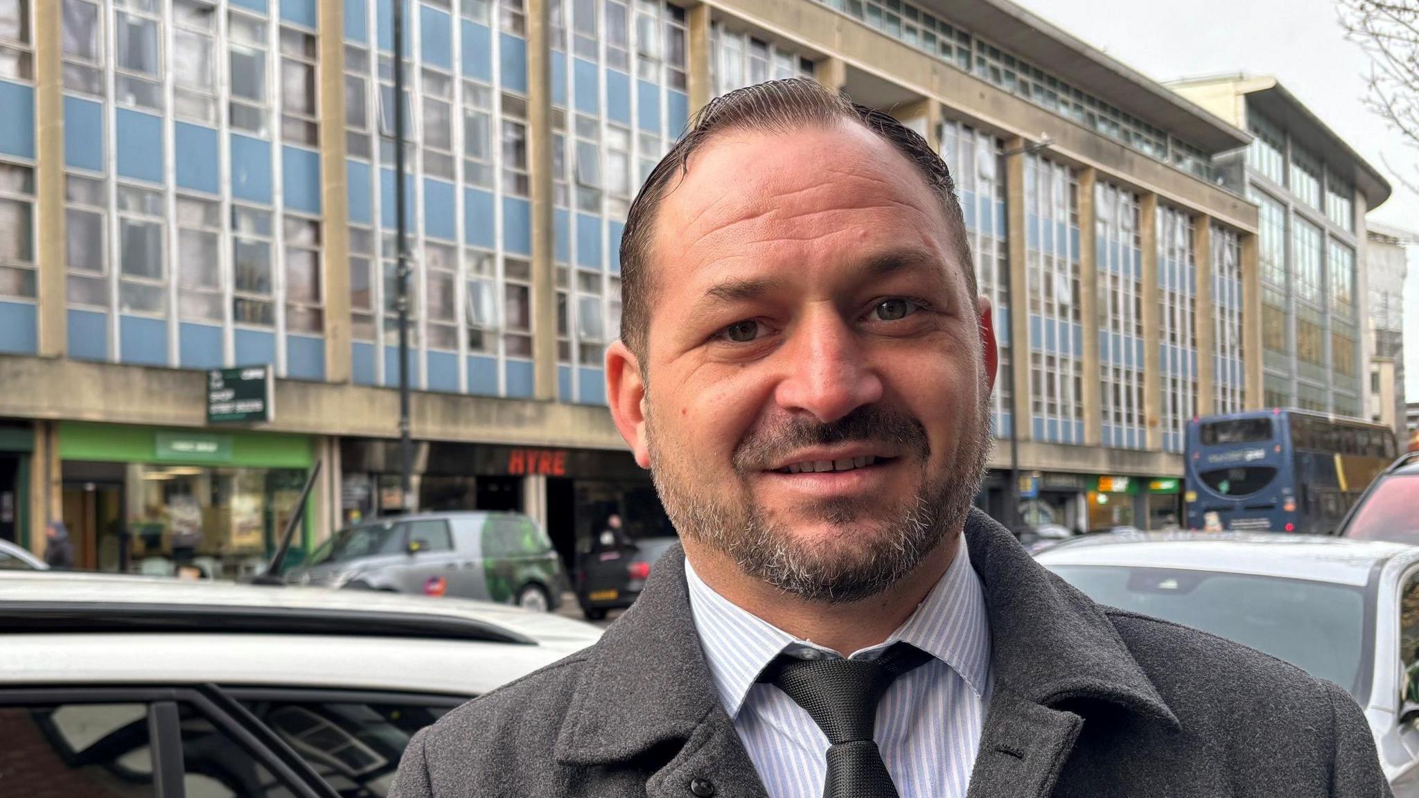 Mr Caraba, smiling at the camera on a busy street. He has dark hair, slicked back, and short grey facial hair. He is wearing a smart grey coat, a light blue and white striped shirt and a grey tie. 