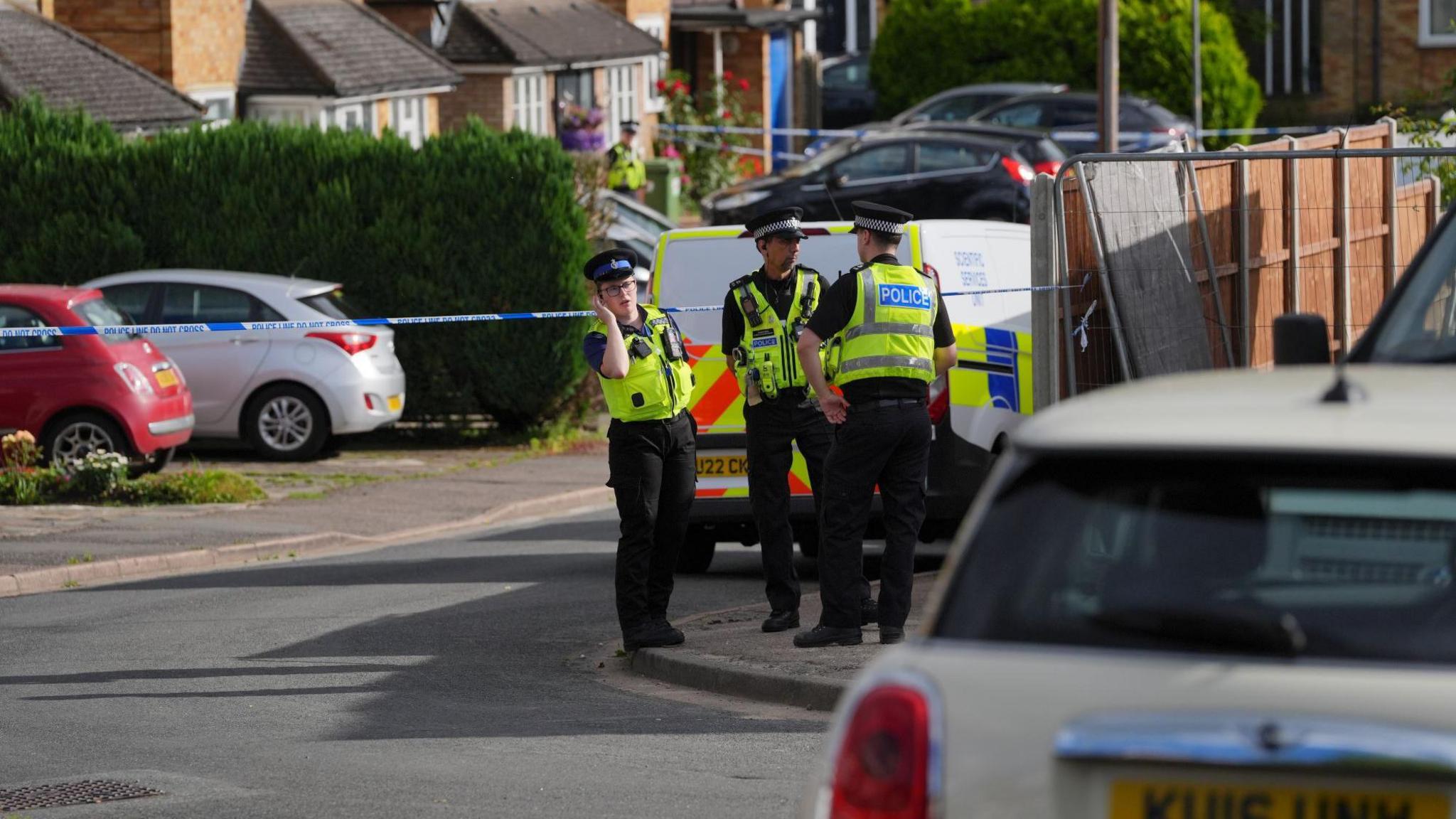 Uniformed police officers are standing on a pavement beside a road. There is blue and white police tap cordoning off a road and terraced houses are in the background.