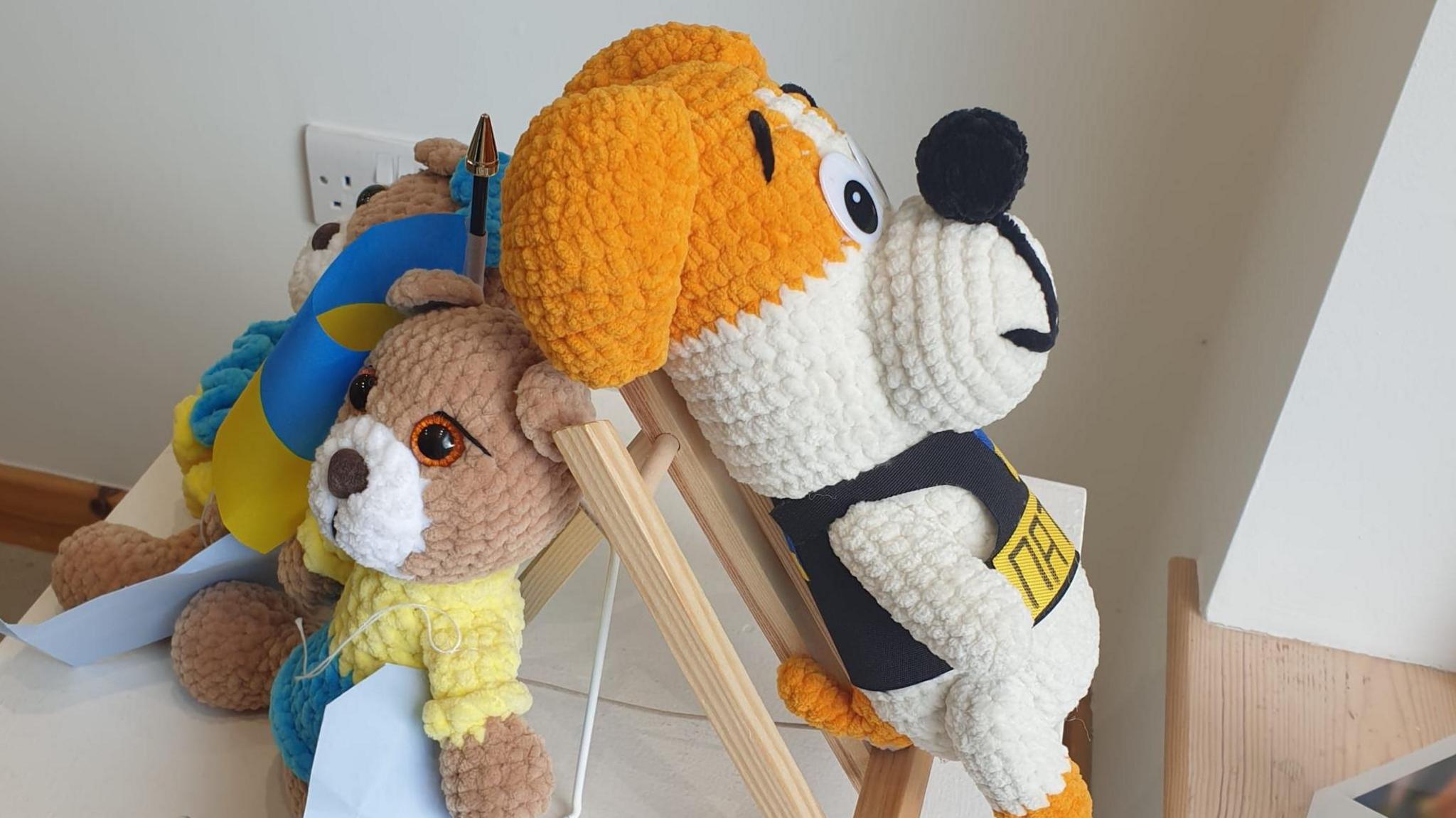 Knitted dolls. One is a dog with a white and orange head. Another is a dog wearing a yellow t-shirt. It has a brown head and a white nose. They are on display on a table.