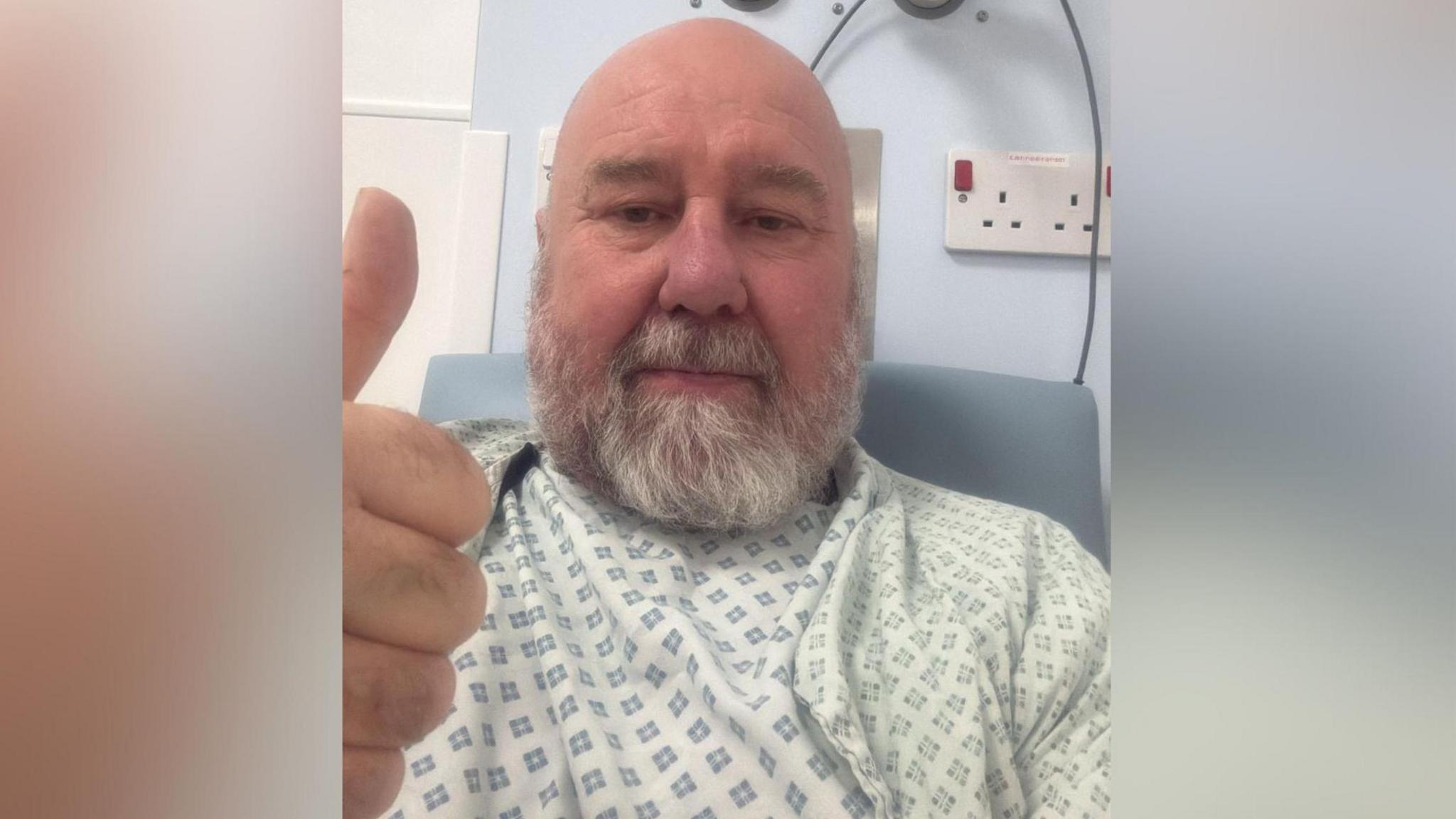 Danny giving a thumbs up to the camera while sitting in hospital. He is wearing a patient's gown.