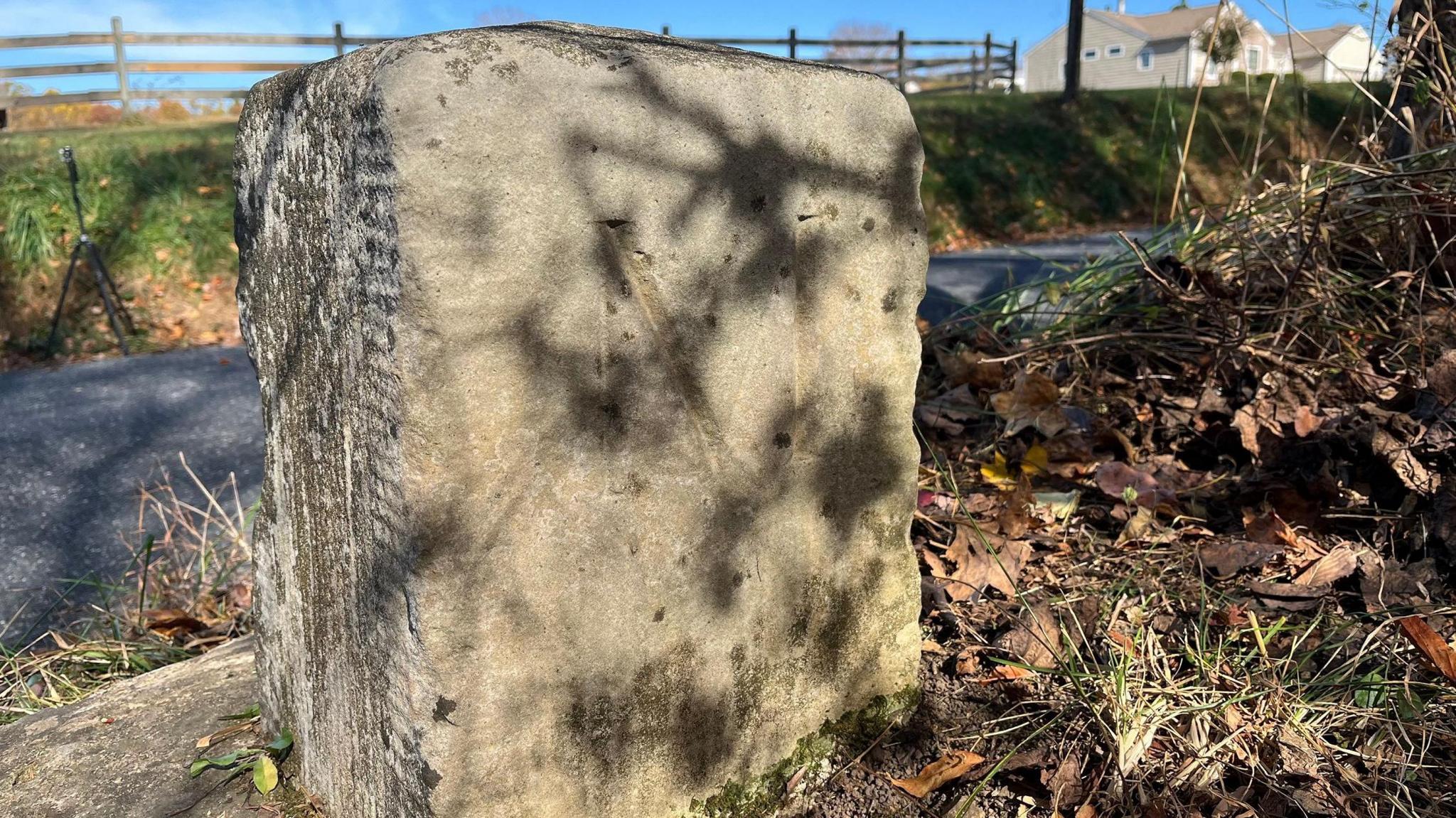 One of the marker stones. It is a low block of stone, set into the earth, ad has clearly been weathered over the years. Despite this, an engraved letter M can be seen written on the stone, which appears to be next to a small road. 