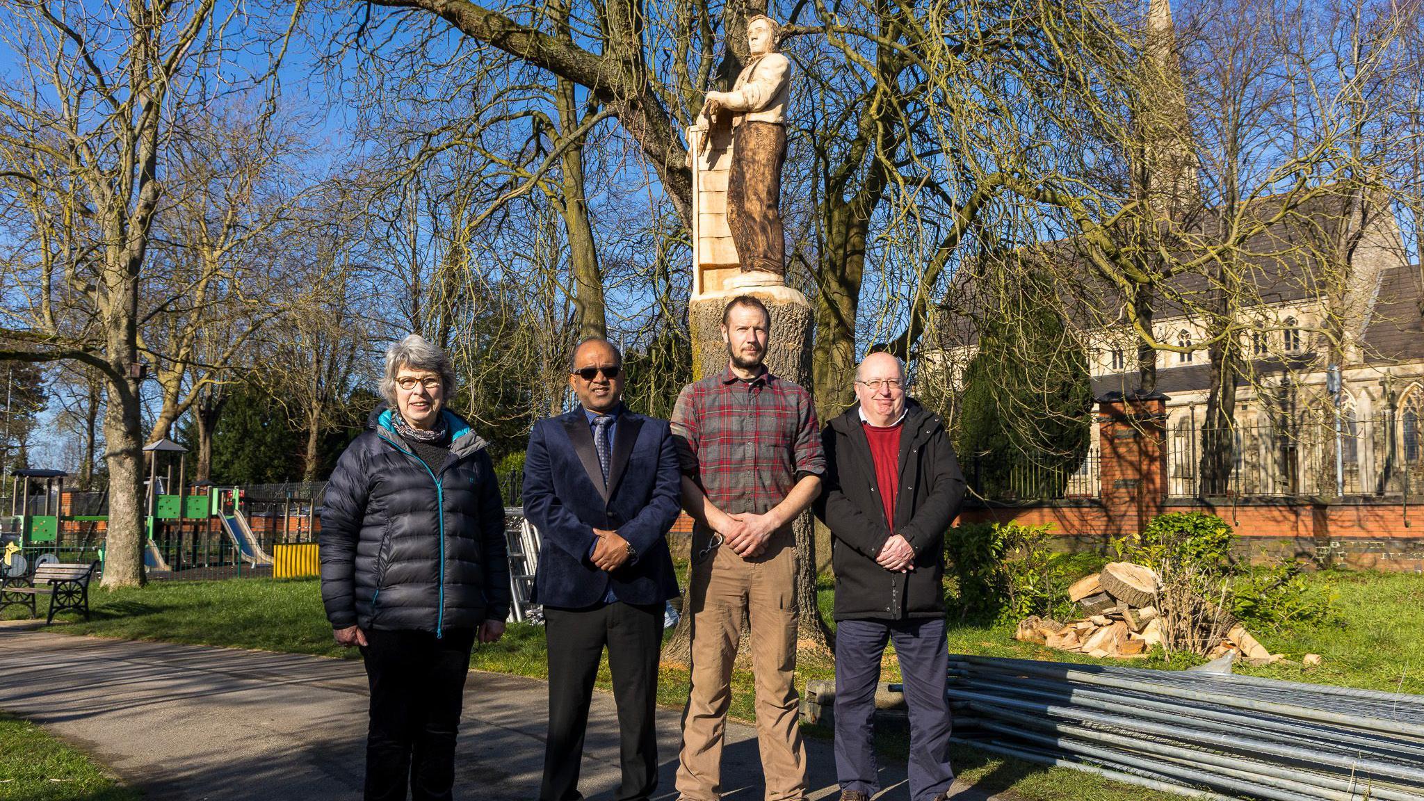 Four councillors (a woman on the left and three men on her right) are standing in front of a wooden statue in a park, on a sunny day.