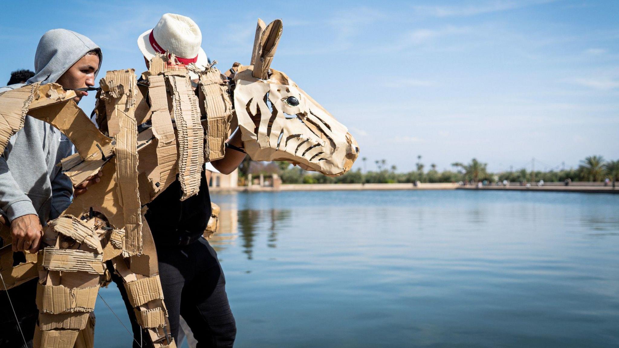 A large cardboard and plywood animal that resembles a zebra, by a body of water in Marrakesh. There are two men behind the puppet, one wearing a grey hoodie and the other in a white hat, holding it in position.