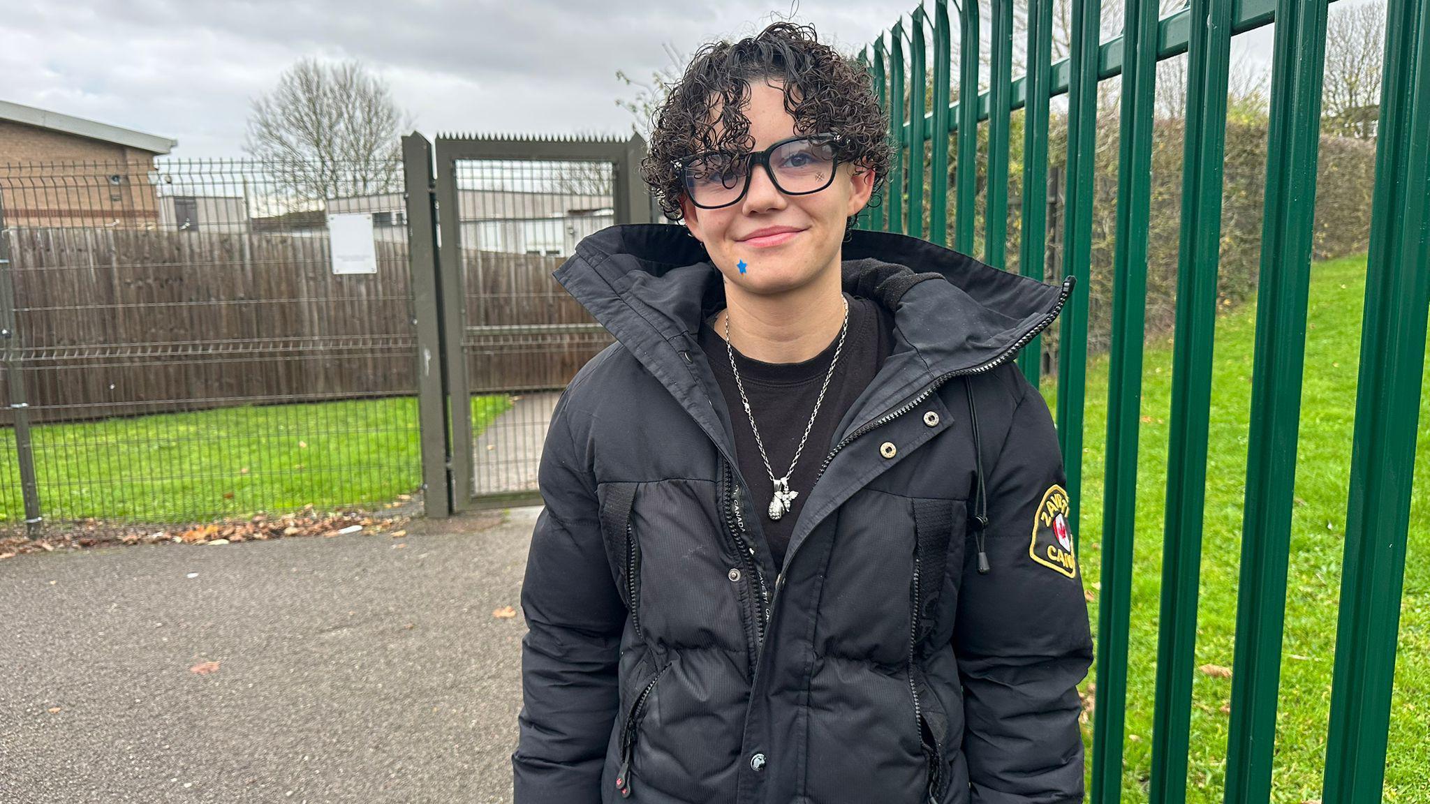 A woman with curly dark hair and a black coat. She is wearing glasses and stood in front of the school gates.