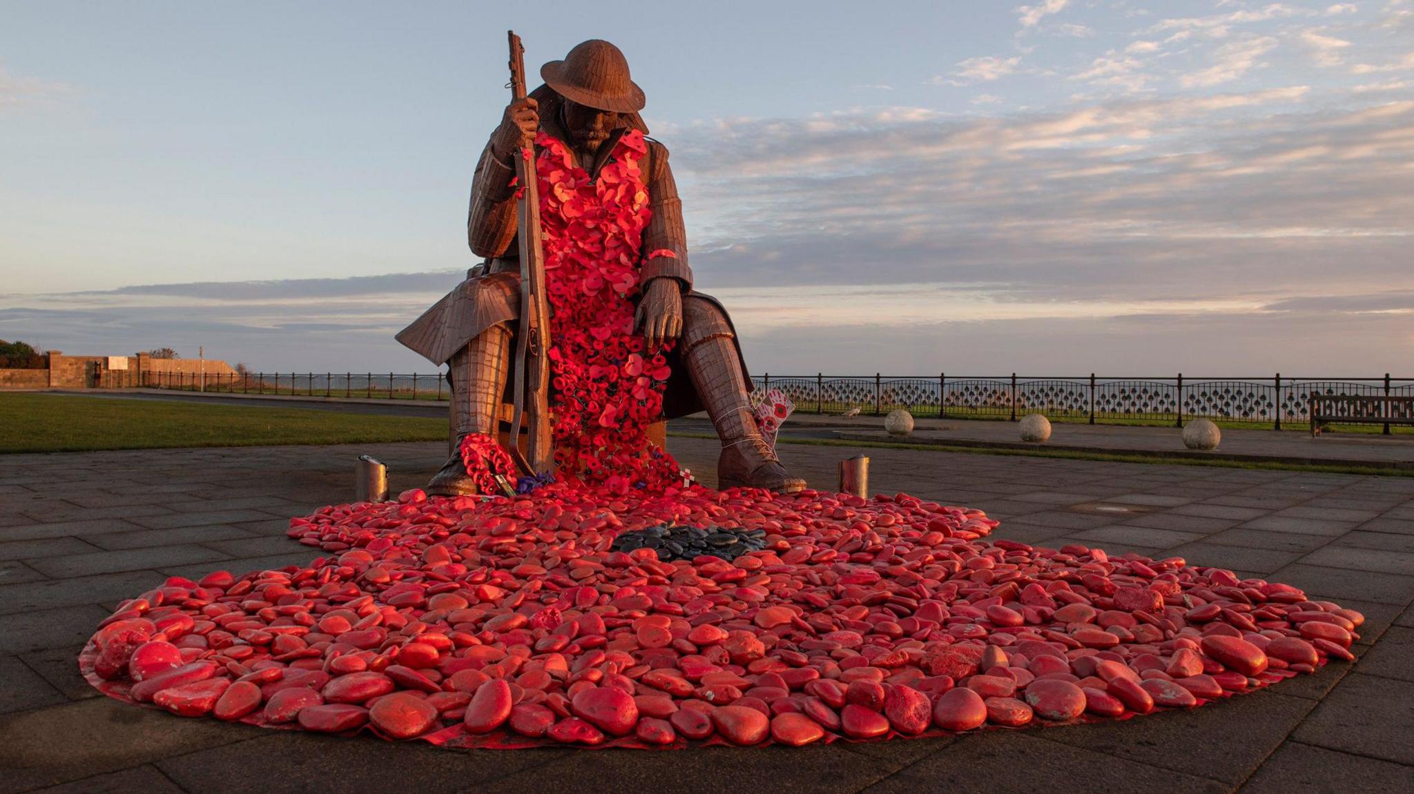 A large statue of a soldier looking down at the ground with a large gun in one of his hands which rests on the ground. It is made of a brown metal and the light appears to be reflecting off of it, giving it an orangey glow. There are poppies draped around the front of the soldier, which cascade down onto the ground where there are many red rocks. The sea can be seen behind the statue and the sky is blue with wispy clouds.