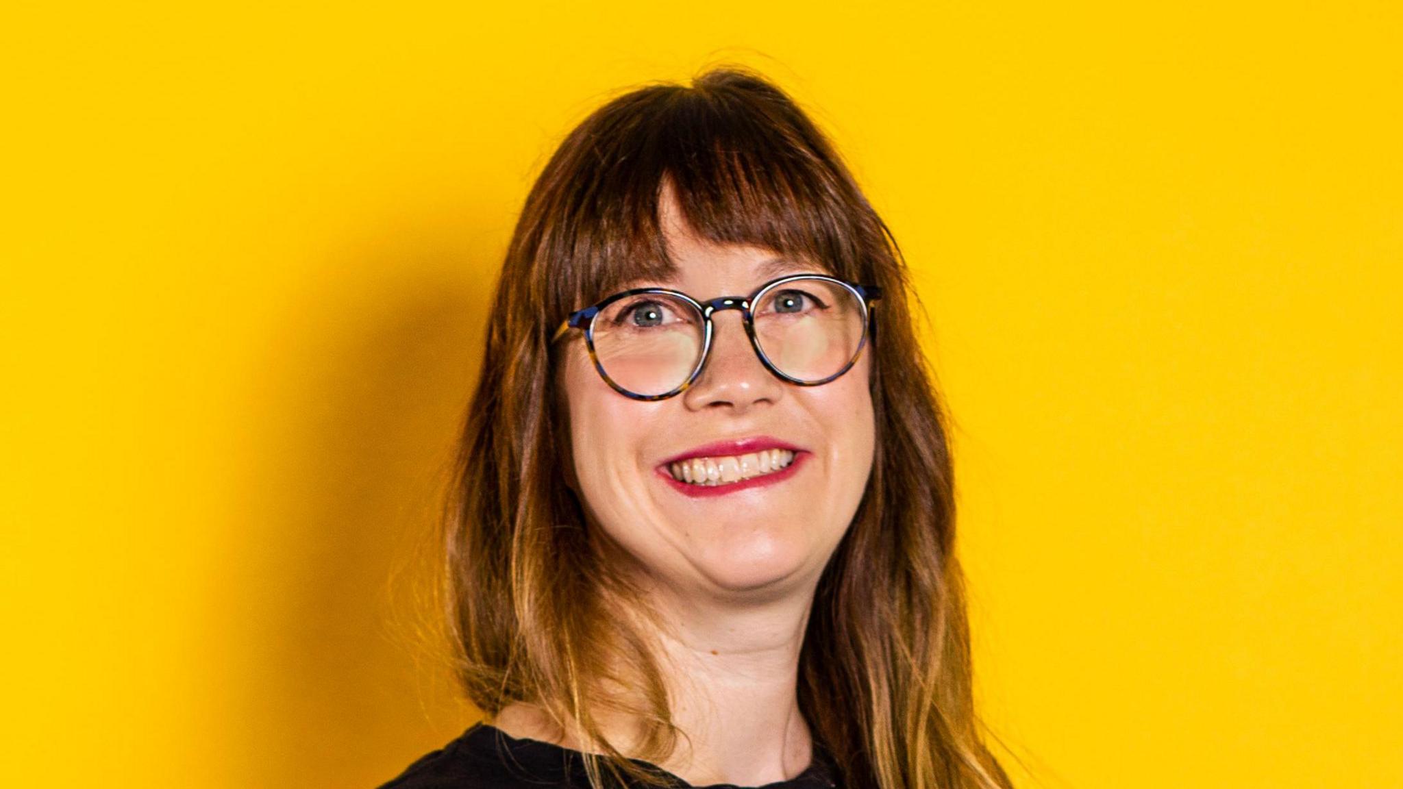 A head and shoulders image of Danielle Ramsey against a yellow backdrop. She is wearing glasses and looking into the camera and smiling.