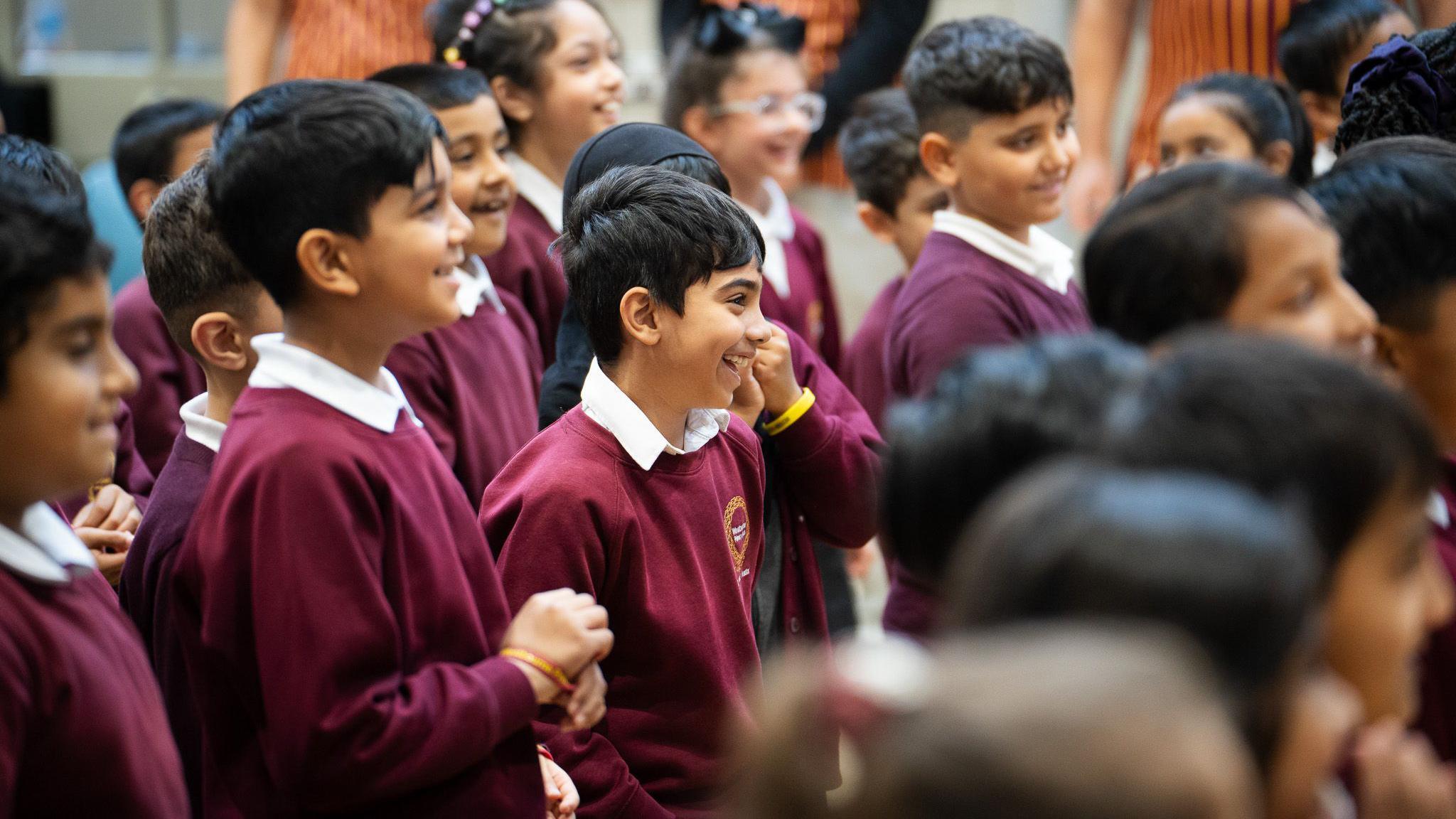 A group of school children standing and smiling during the workshop. They are wearing burgundy sweatshirts and white polo shirts.