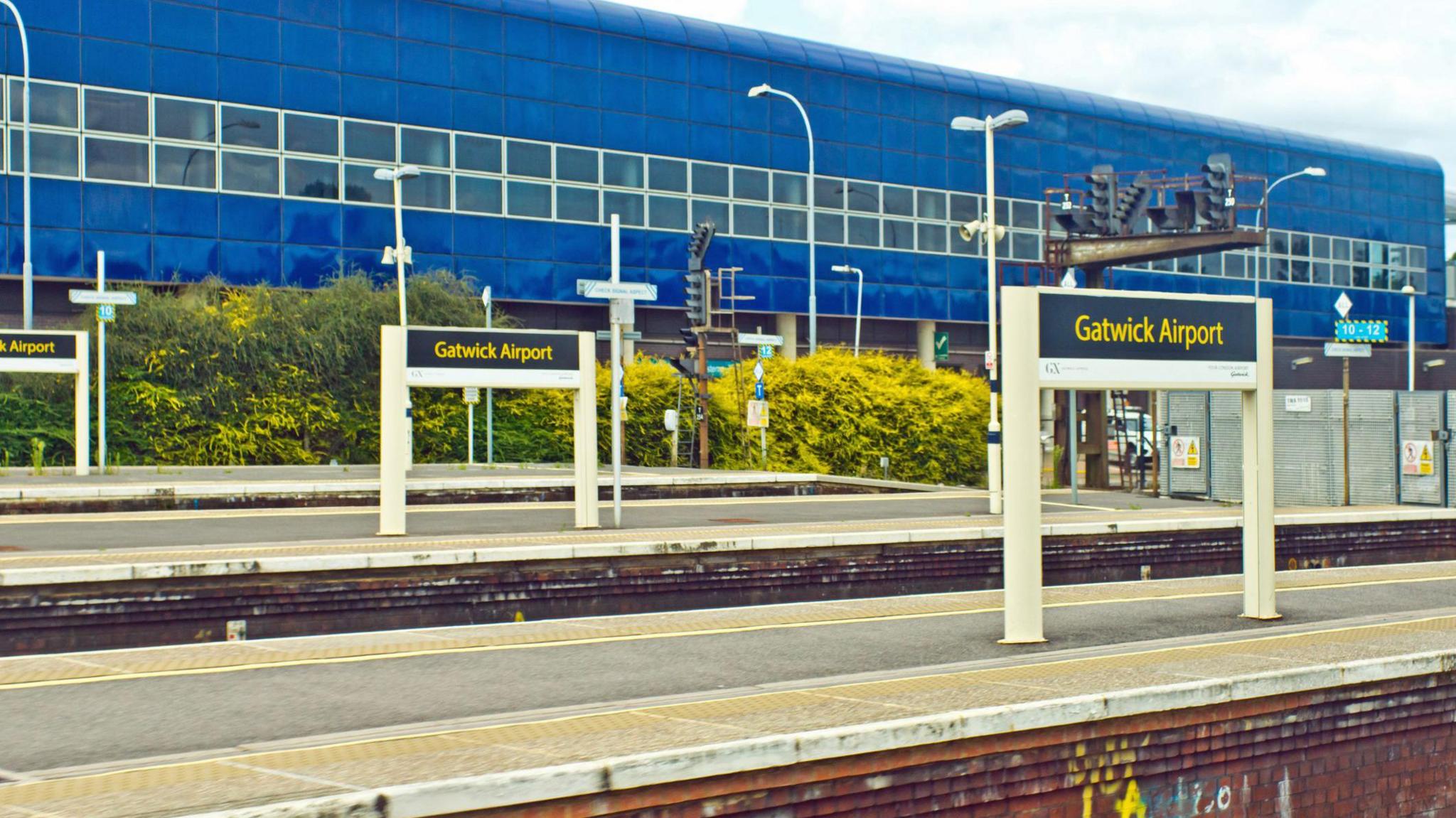 Gatwick Airport train station - stock photo. A concrete platform, with three signs for Gatwick Airport. There are street lamps and a large blue building in the background