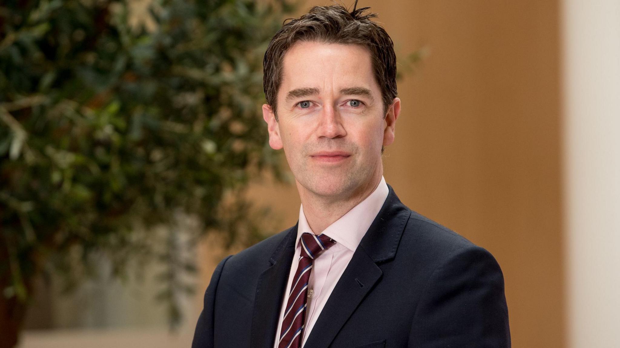 Man with short dark hair and clean shaven, wearing navy suit, light shirt and burgundy striped tie