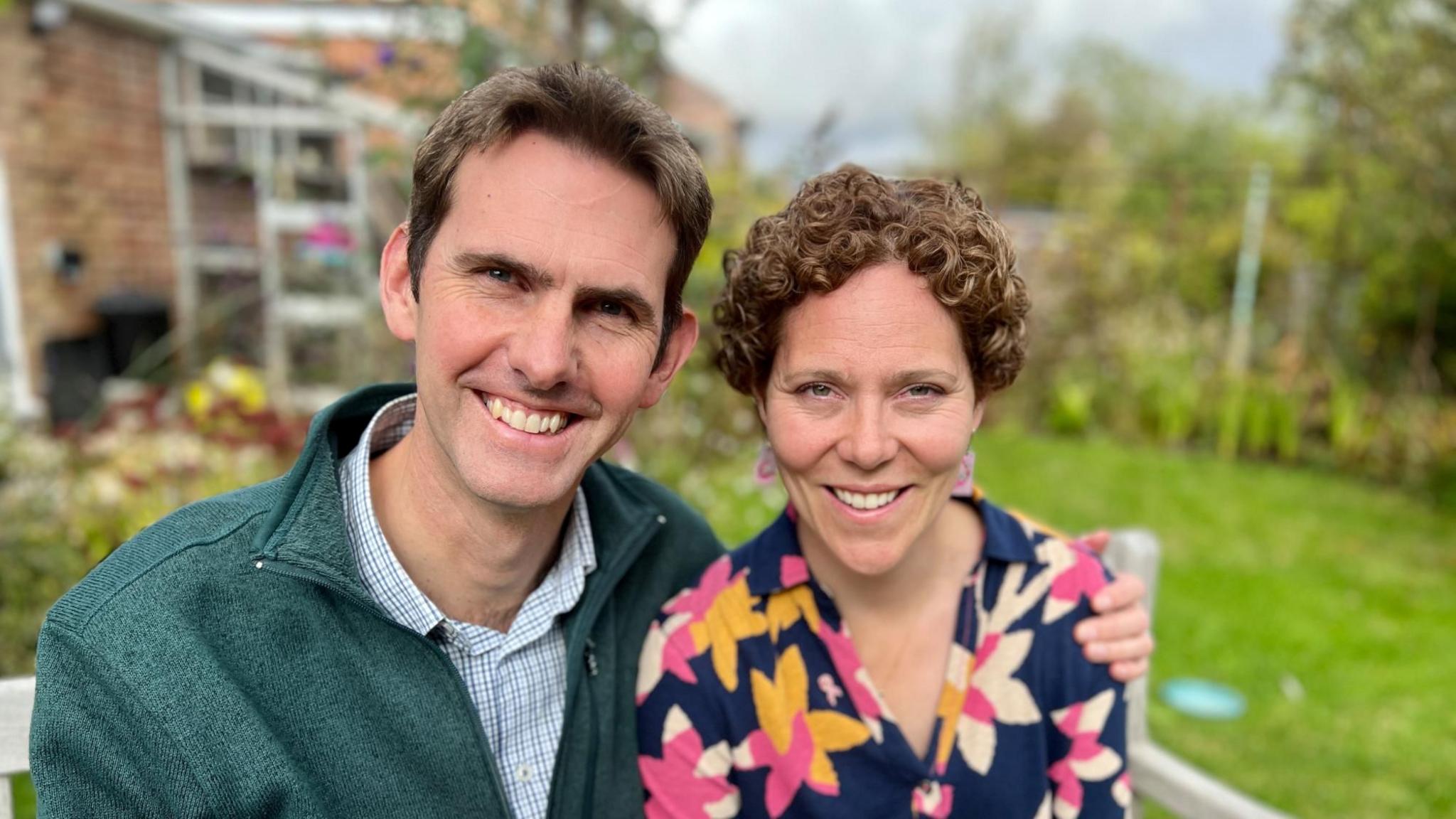 Julia Beeden sits with her husband in her garden in Cambridgeshire. Julia is wearing a flowery navy top, and has curly hair that has grown back after chemotherapy and her husband is wearing a green fleece with a checked shirt underneath. They are both smiling.