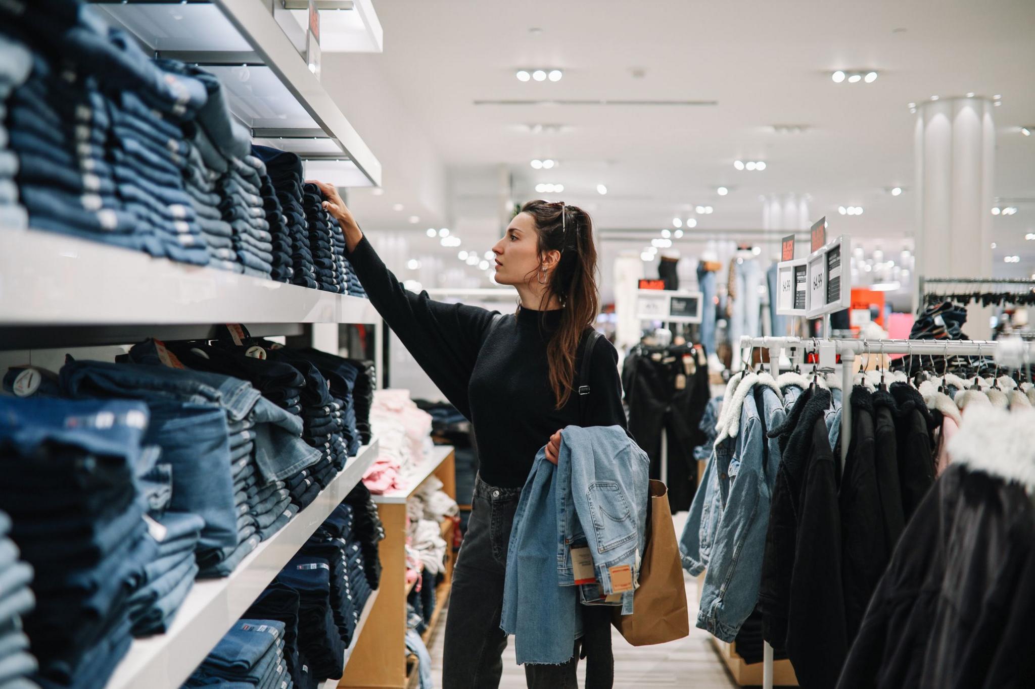 Young woman with brown hair in clothing store, looking at shelves stacked with jeans.