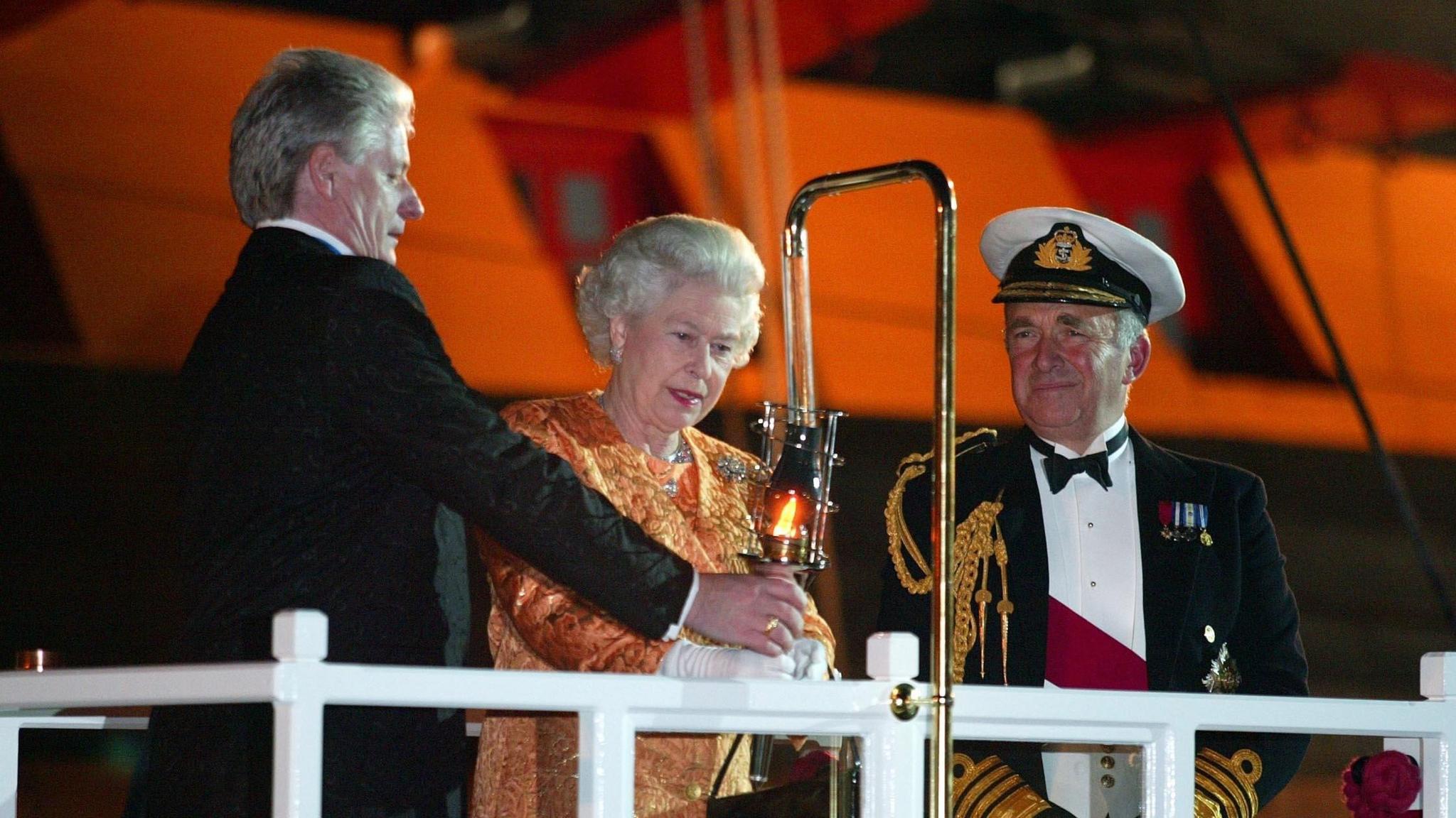A man dressed in a black suit helps the Queen to light a beacon with a flame. She's wearing an orange and gold outfit, and a man wearing a uniform is standing next to them watching.