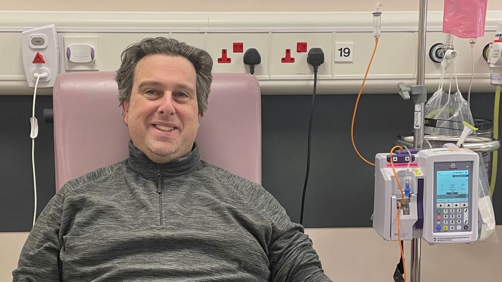 A man in a charcoal grey quarter zip top sits in a chair in a hospital. There are pieces of medical equipment surrounding him. 